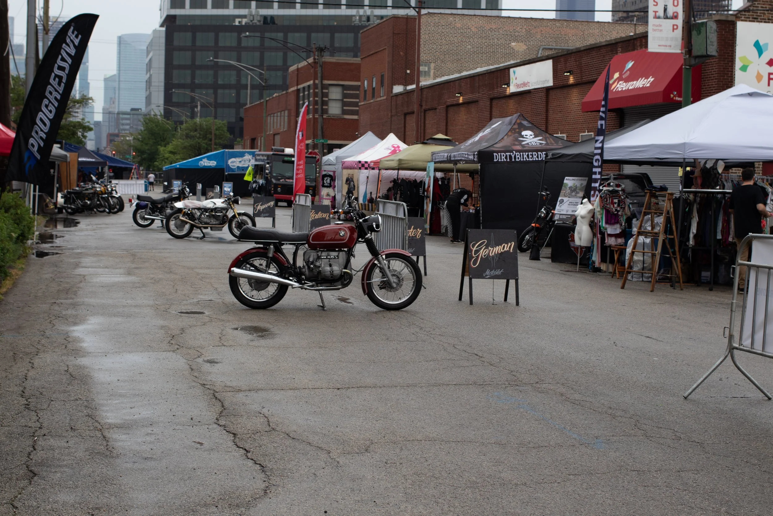 Outdoor street fair with tents and motorcycles, in an urban area with tall buildings and a wet pavement.