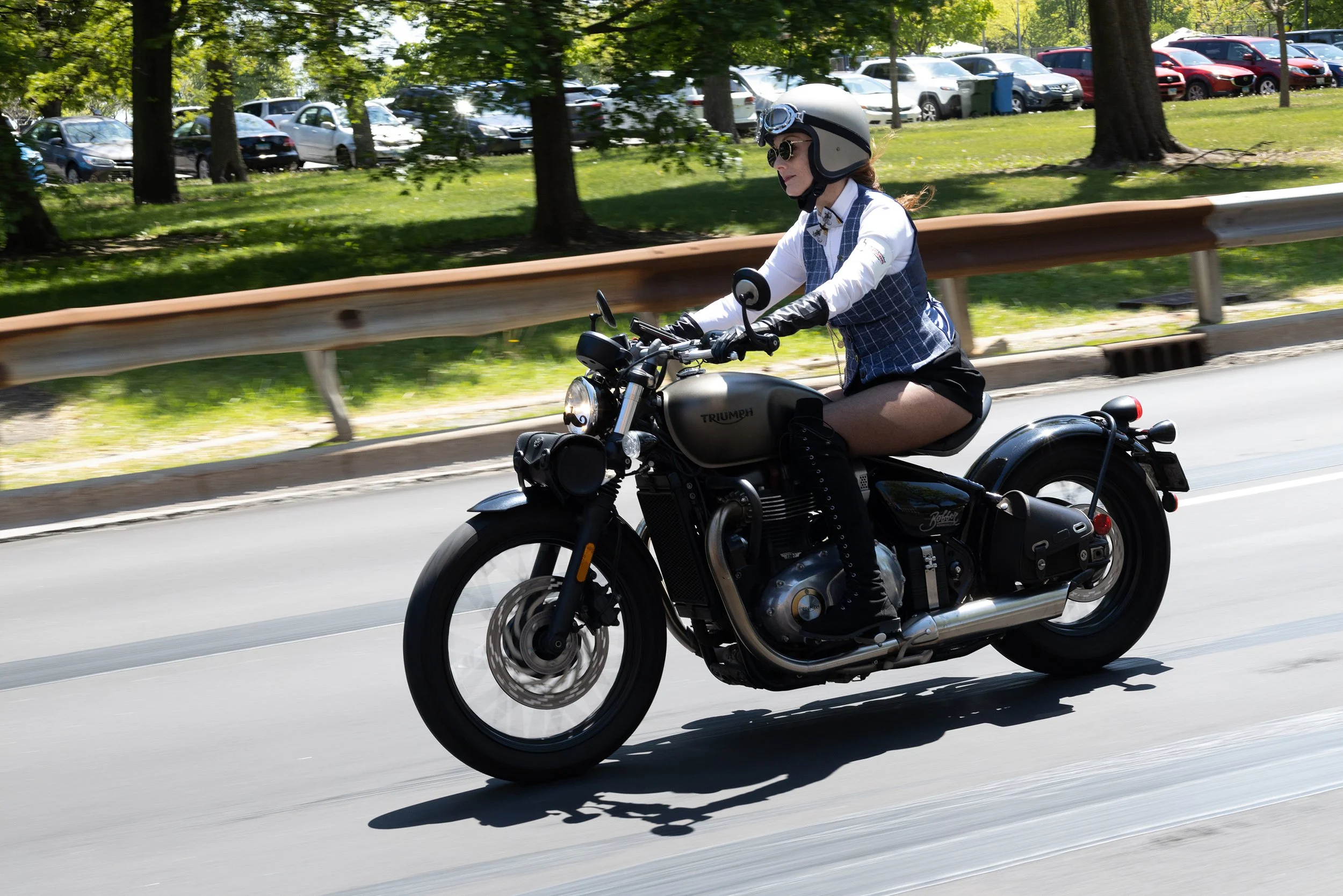 A woman riding a Triumph motorcycle on a road near a park with trees and a parking lot in the background.