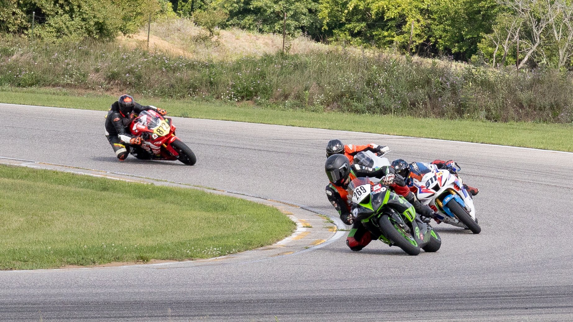 Motorcycle racers lean into a curve during a race on a paved track surrounded by green grass and trees.