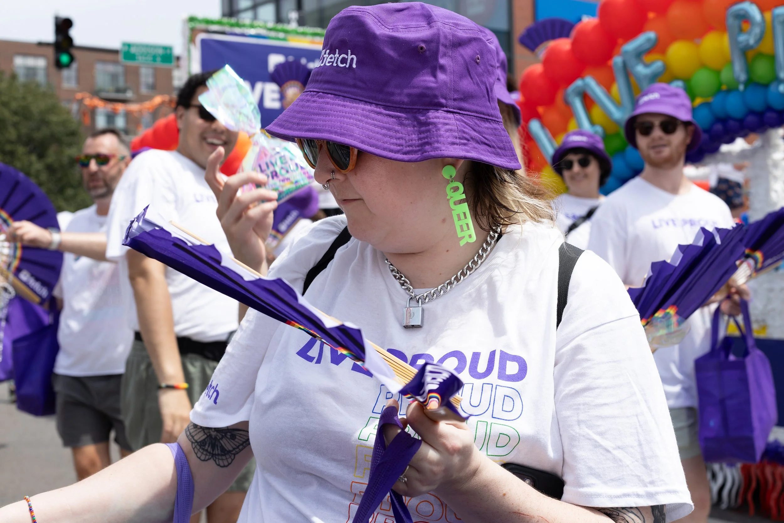 People participating in a pride parade or event, wearing white t-shirts with colorful text, purple hats, and accessories with rainbow colors. One person in the foreground is attaching a purple ribbon, while others in the background are smiling, weari