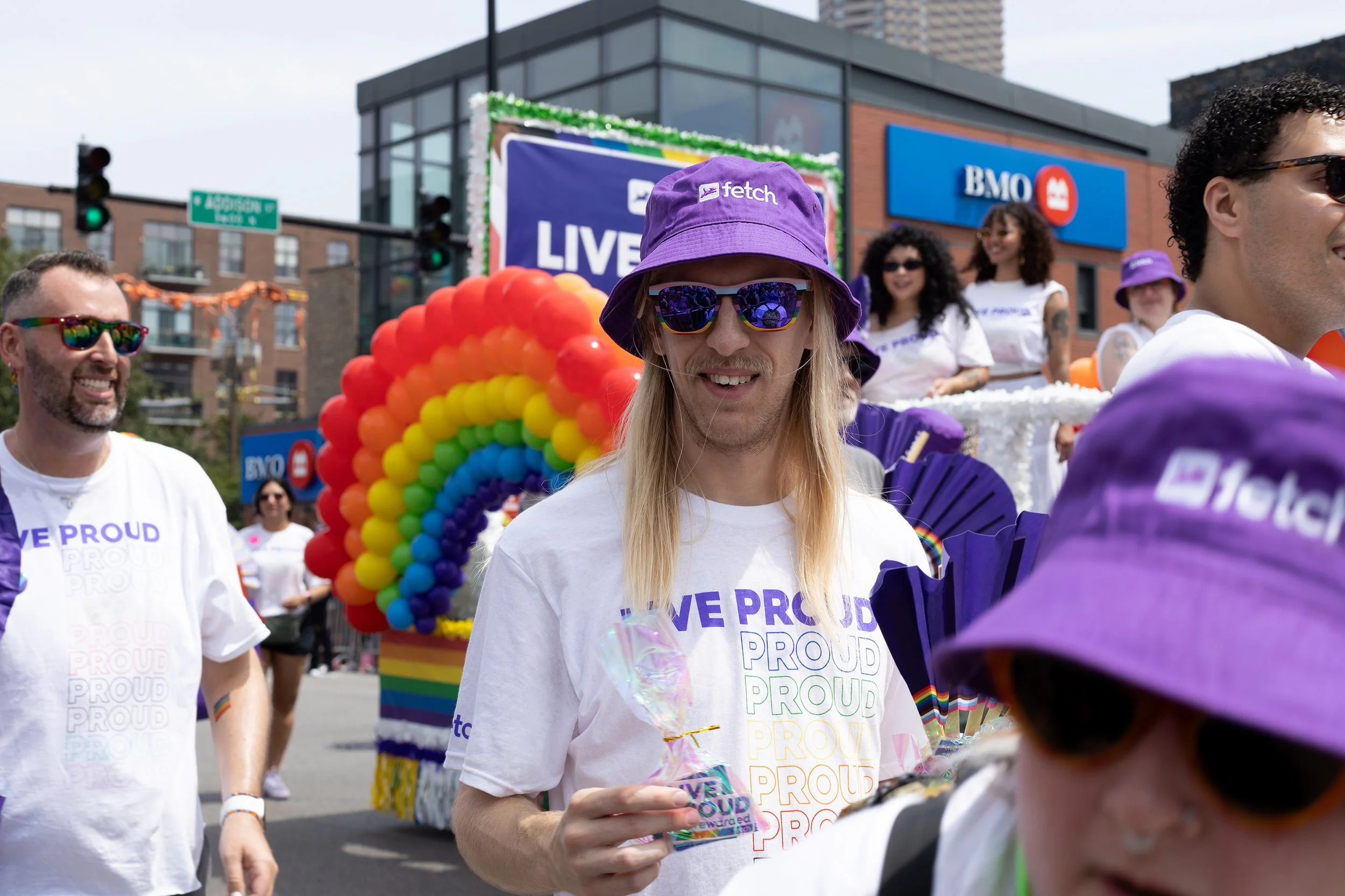 Participants in a pride parade wearing white t-shirts with rainbow text and accessories, including purple hats and sunglasses, walking past a float decorated with rainbow-colored balloons and a sign that reads 'LIVE PROUD' in a city street.
