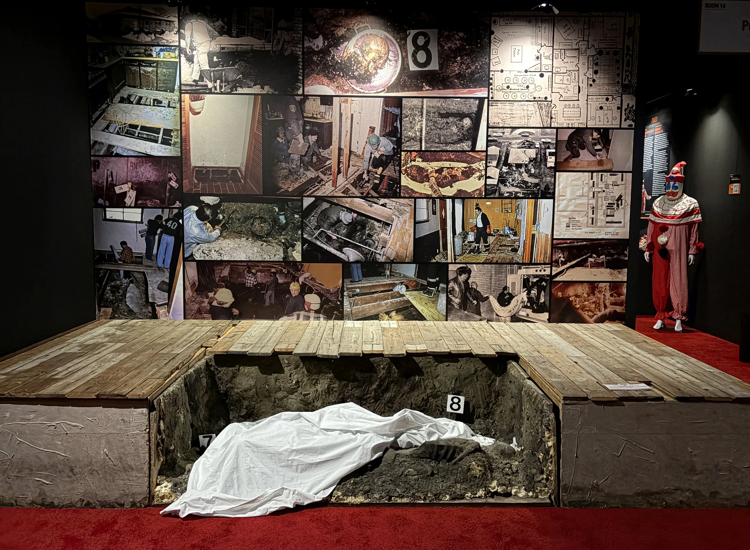 Exhibition display with multiple photographs of construction workers, architectural plans, and excavation site beneath a partially removed floor, with a clown statue dressed as Pennywise from Stephen King's 'It' to the right.