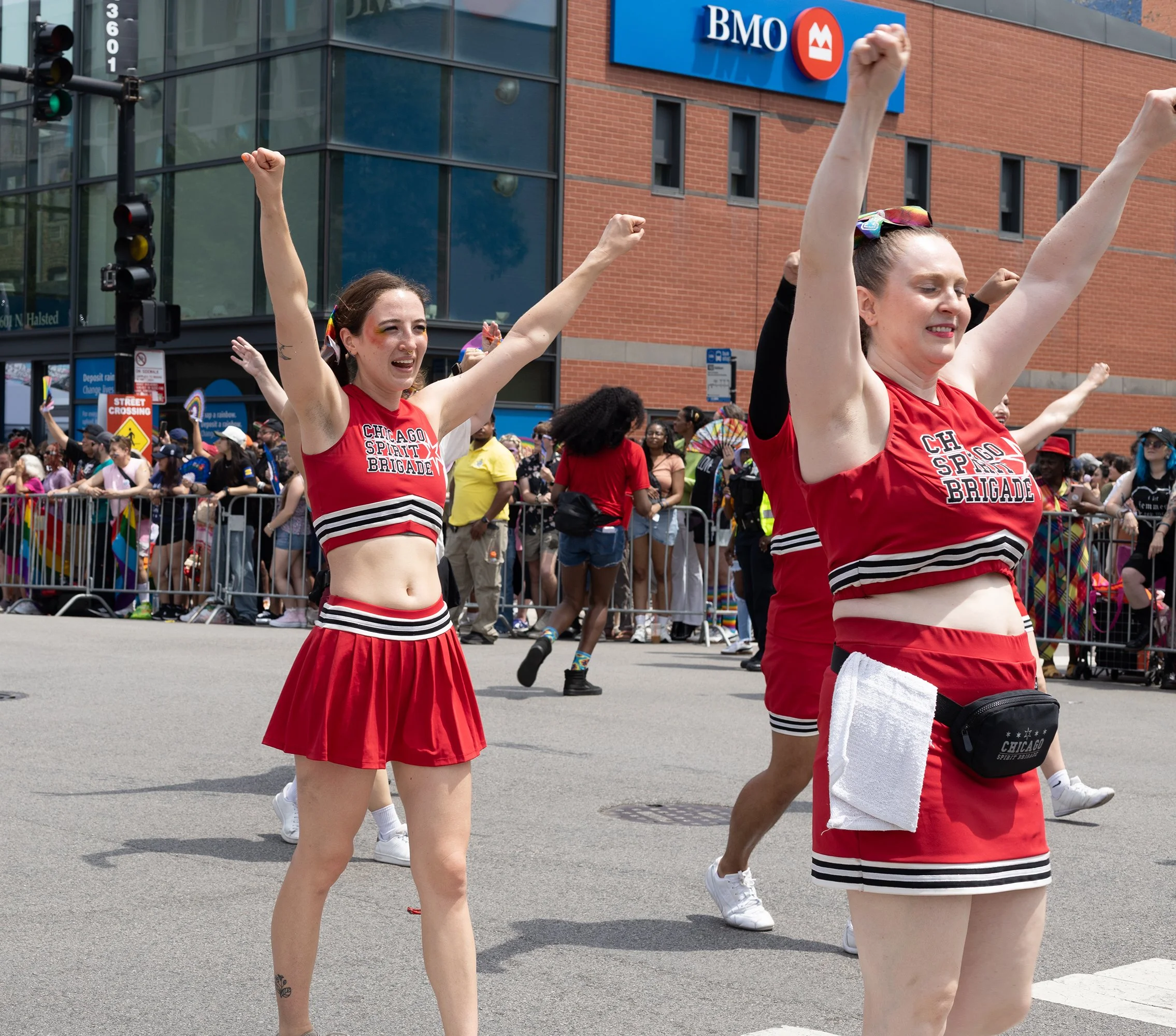 Two women wearing red cheerleading uniforms with 'Chicago Spirit Brigade' written on them, participating in a parade or public event in an urban setting with a crowd and a building with a BMO sign in the background.