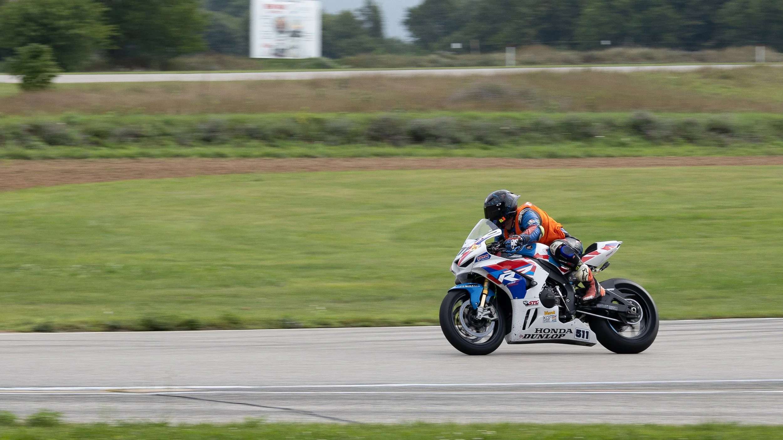 A motorcyclist riding a white, red, and blue Honda motorcycle on a road, wearing a black helmet and racing gear with an orange vest, with green grass and trees in the background.