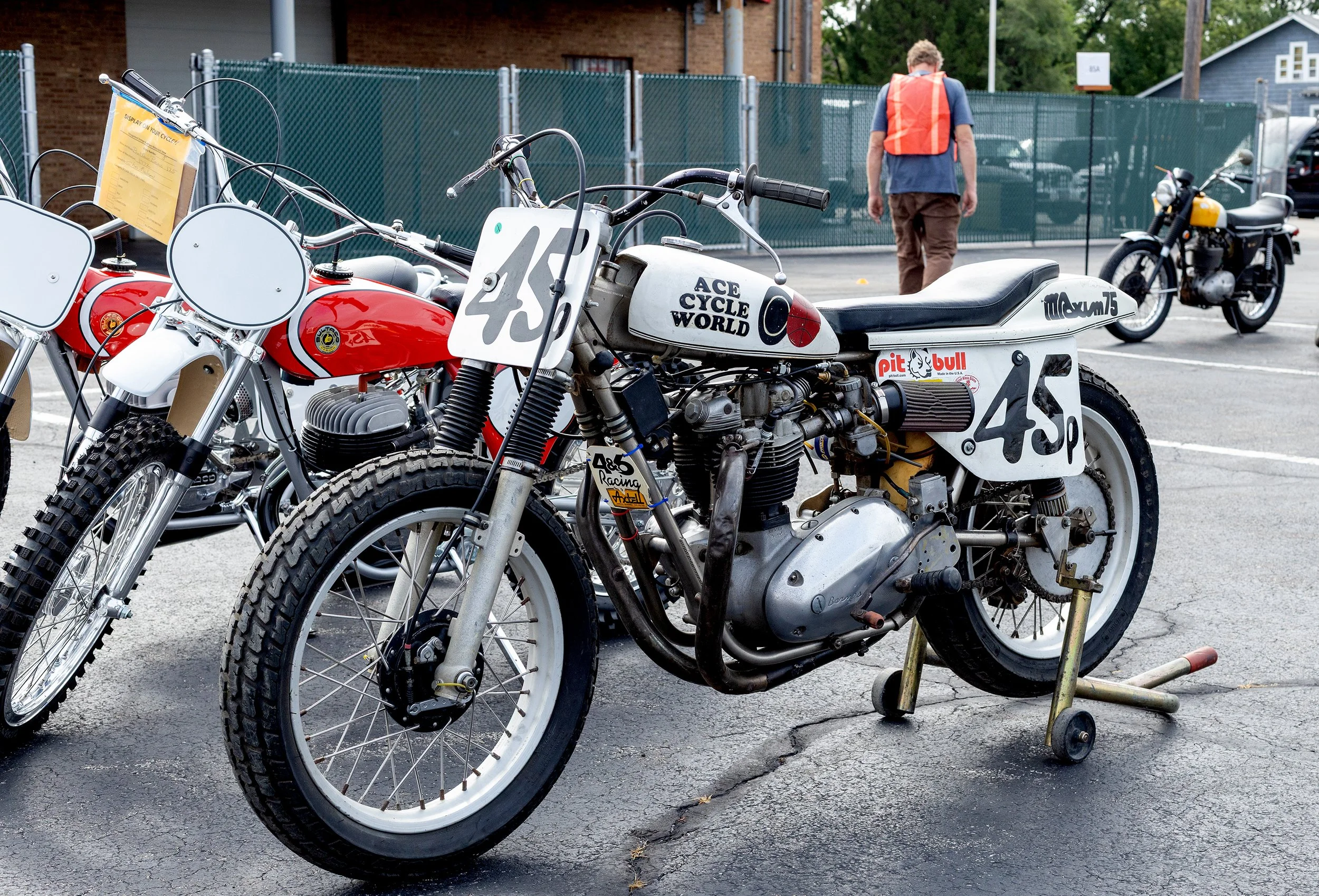 A row of vintage race motorcycles parked outdoors, with a person wearing an orange safety vest walking in the background near a green fence.
