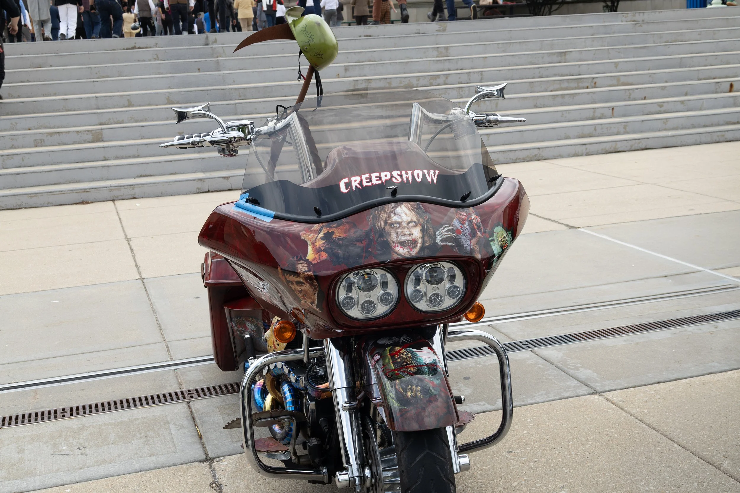 A motorcycle with horror-themed artwork on the front and windshield, featuring a creepy clown face and the word 'CREEPSHOW', parked in front of a set of concrete steps with a crowd of people in the background.