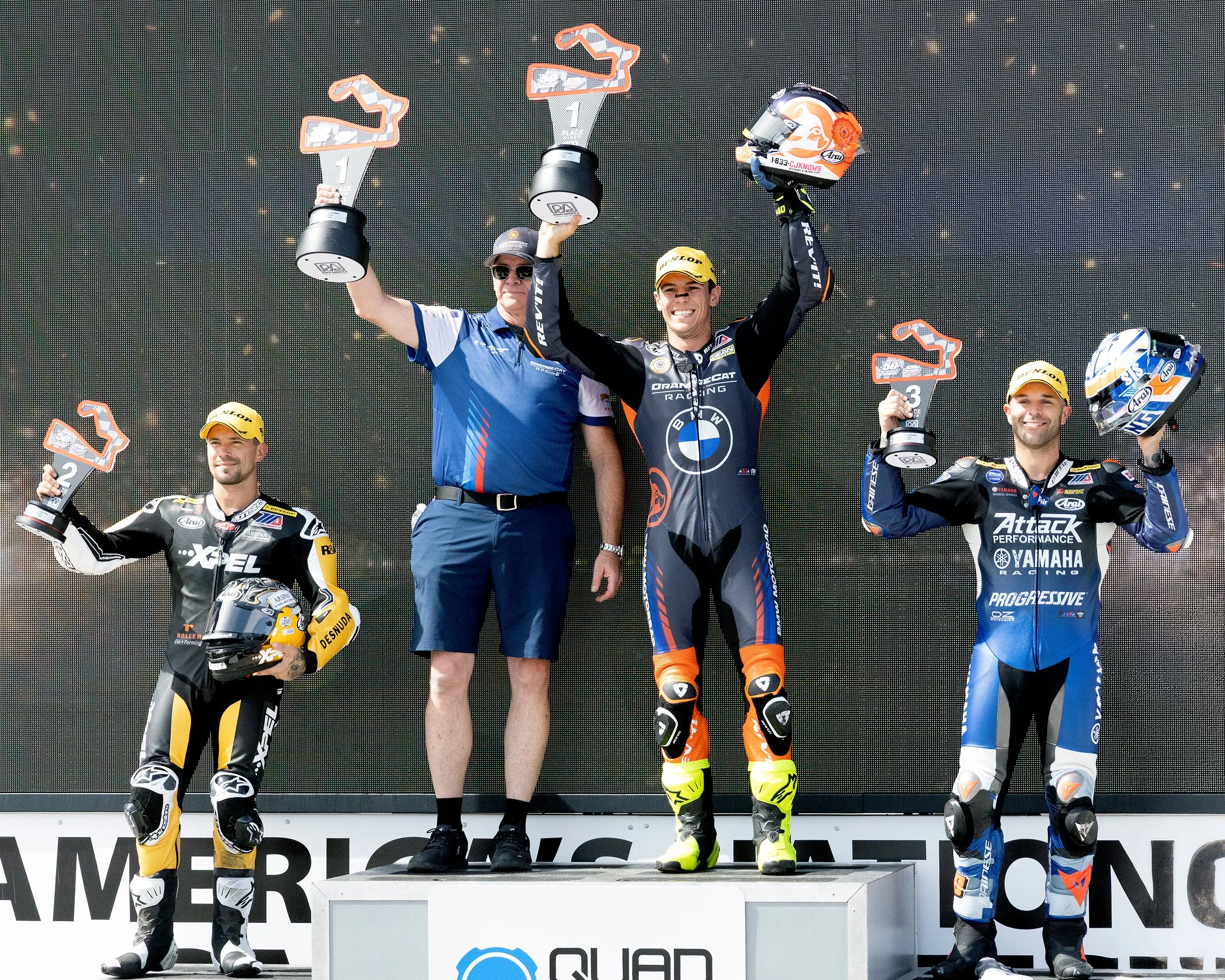 Motorsport winners on a podium holding trophies, with racers in racing suits and helmets, celebrating after a race.