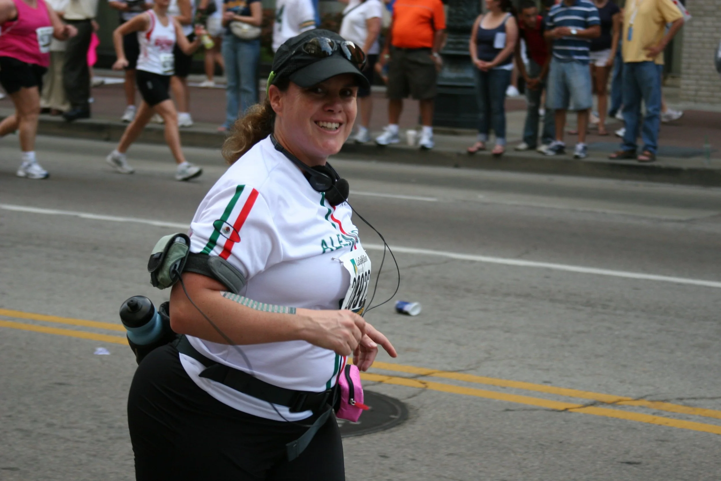 A smiling woman participating in a marathon or running event, wearing a white shirt with Italian flag stripes on the sleeve, a black cap, and audio headphones, with a group of people in the background on a city street.