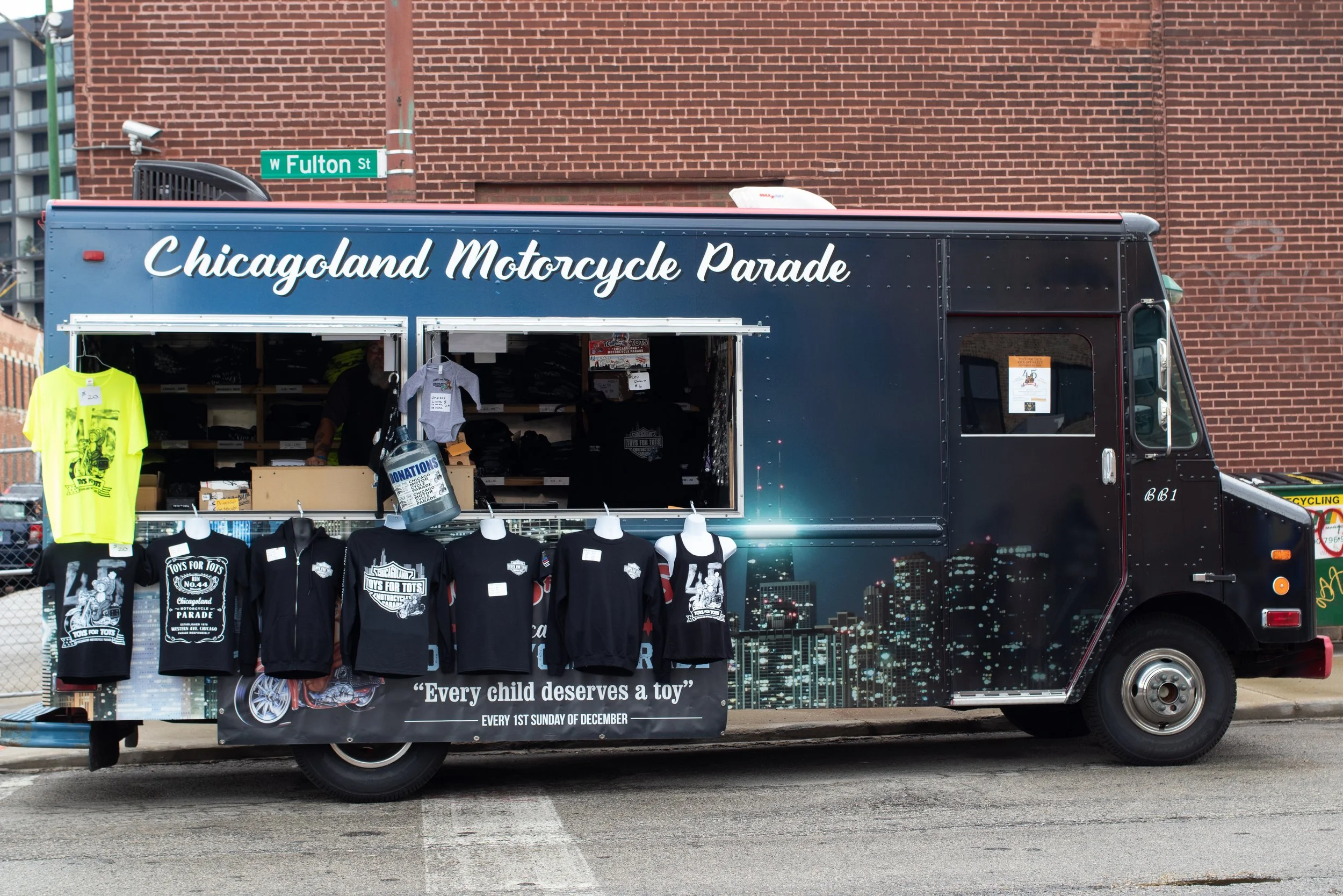 A food truck with a sign that reads 'Chicagoland Motorcycle Parade' parked on a street, with merchandise including T-shirts and clothing displayed outside. There is a banner stating 'Every child deserves a toy' and an event date of the first Sunday o