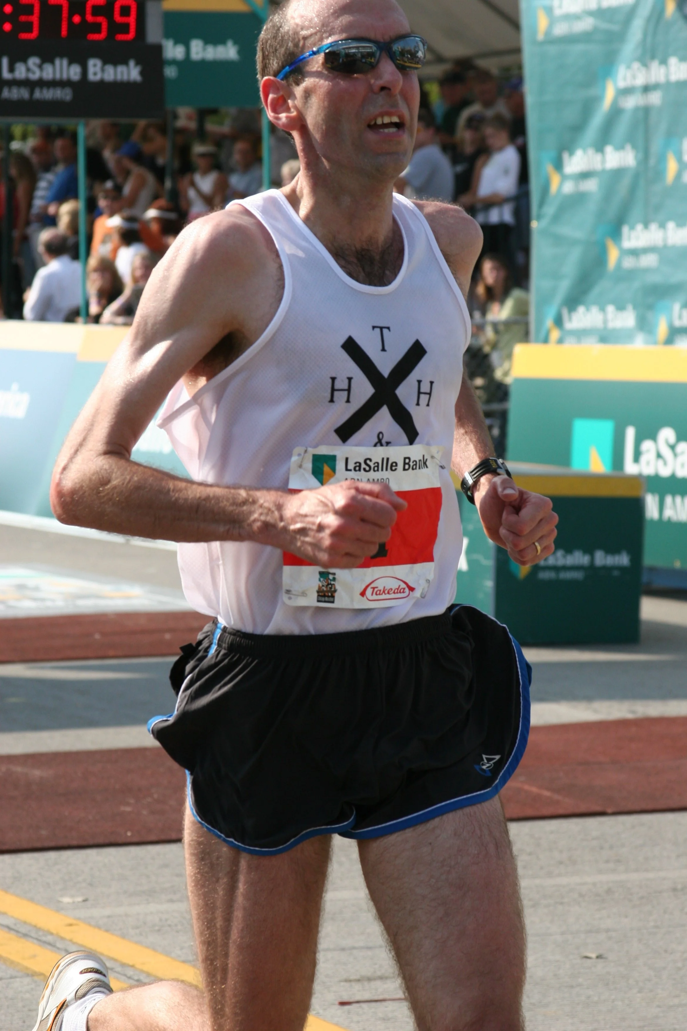 Male marathon runner wearing sunglasses, white tank top, black shorts, running outdoors at a race event with spectators in the background.