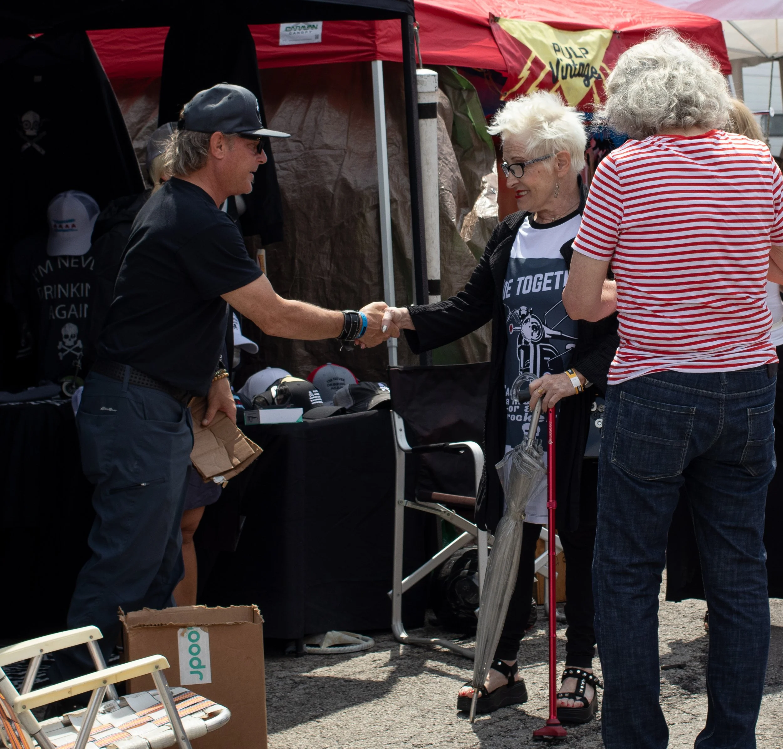 People at an outdoor event exchange greetings, with a man and woman shaking hands under a red canopy, others walking by, souvenir hats and items displayed on tables.