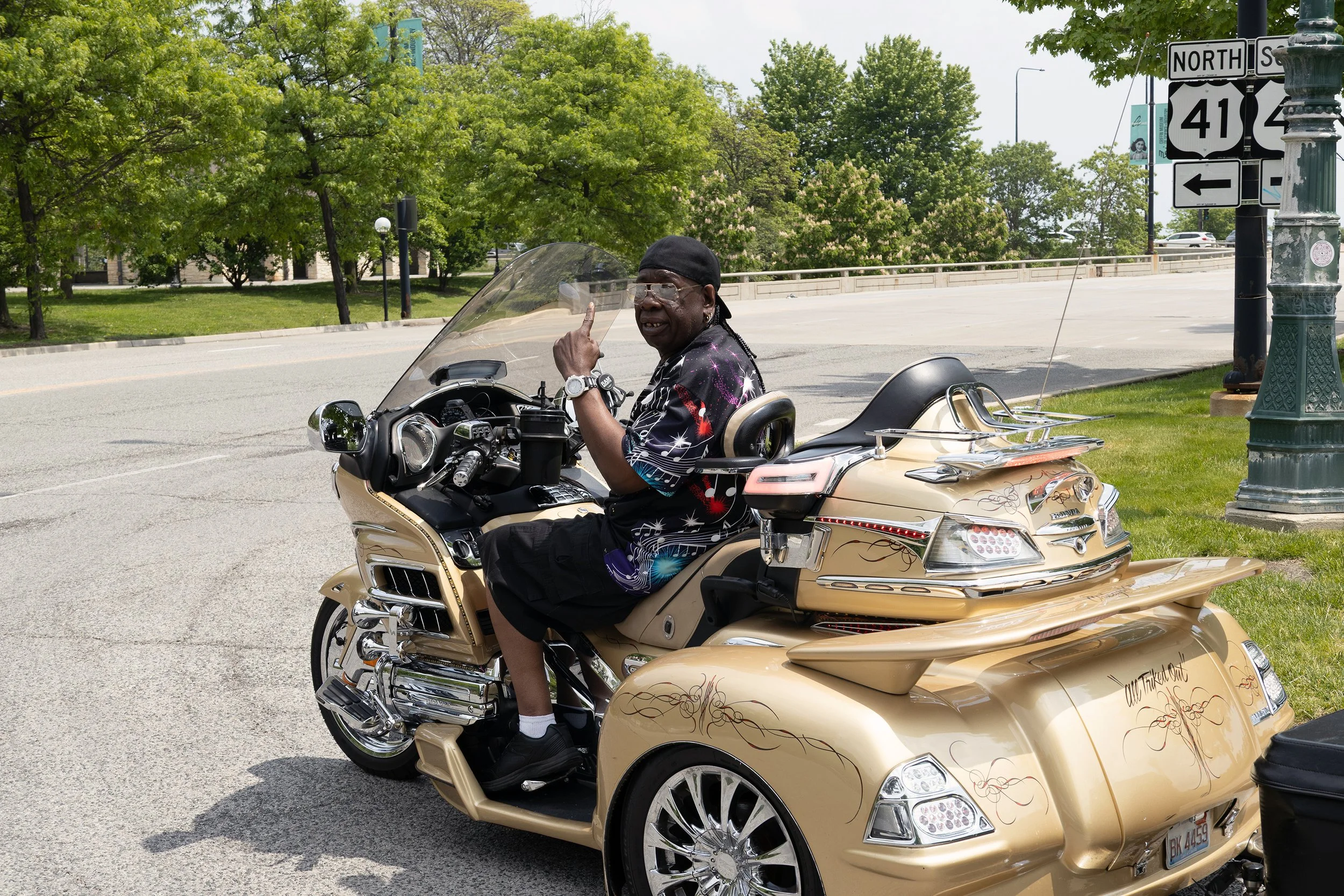 A man wearing glasses, a black head covering, and a colorful shirt is sitting on a gold-colored trike motorcycle, making a peace sign with his right hand, parked near a curb on a street with greenery and traffic signs.