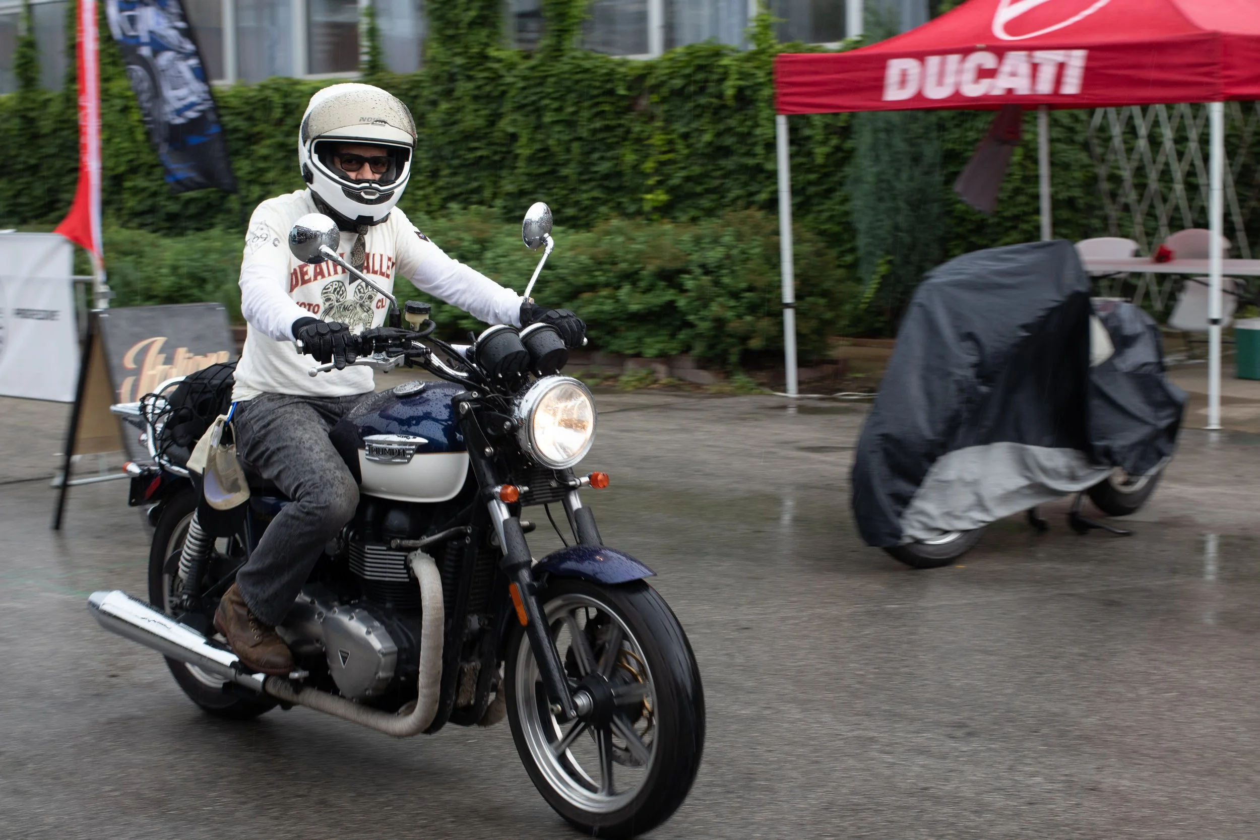 A man wearing a white helmet and sunglasses riding a black motorcycle on a street. He is dressed in a white shirt with a graphic design and dark pants. The background includes a red tent with the word "Ducati" on it, green foliage, and some outdoor f