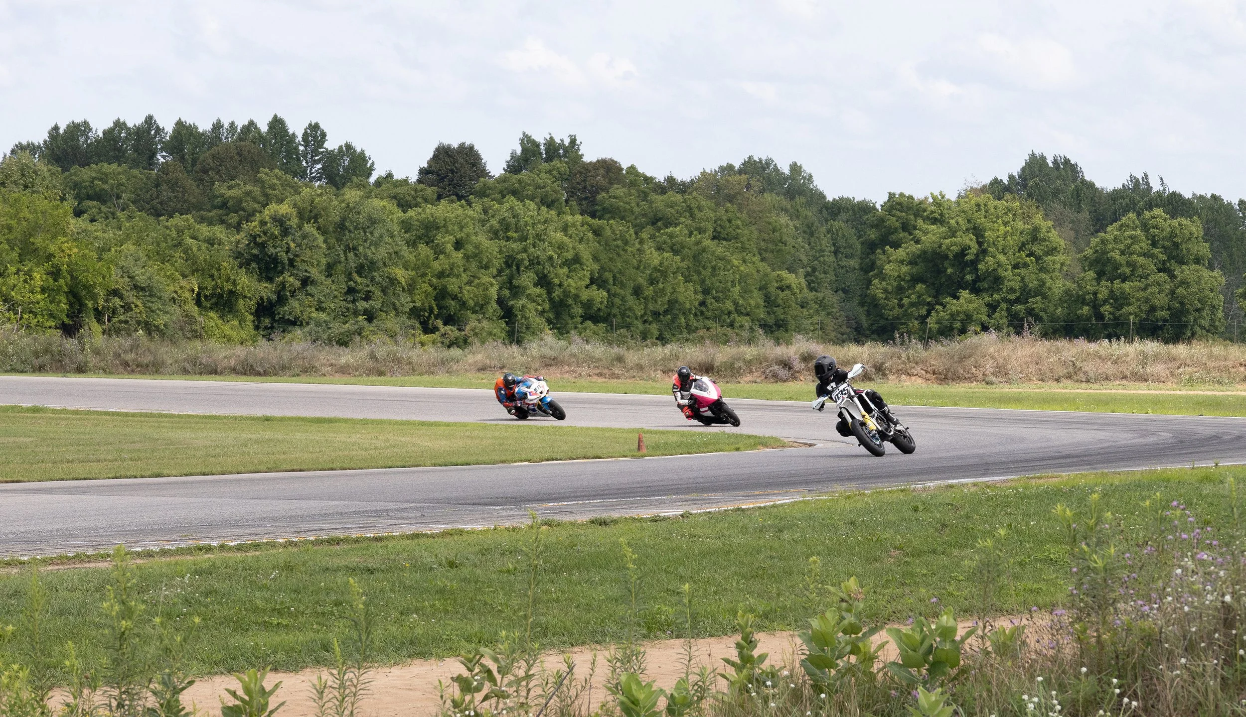 Three motorcyclists lean into a turn on a racetrack, surrounded by trees and grass.