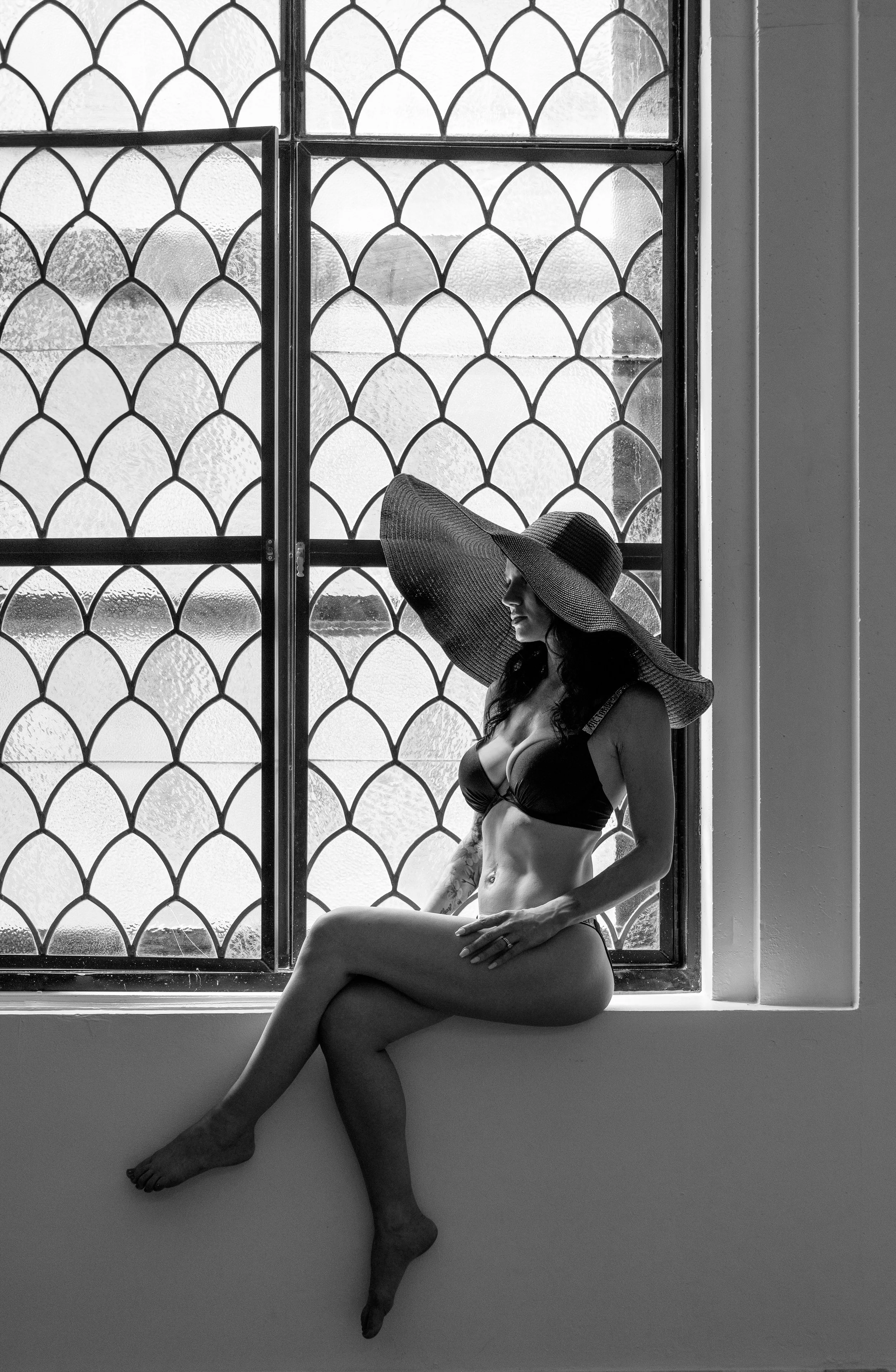 A woman sitting on a window ledge wearing lingerie and a wide-brimmed hat, with a patterned window in the background.