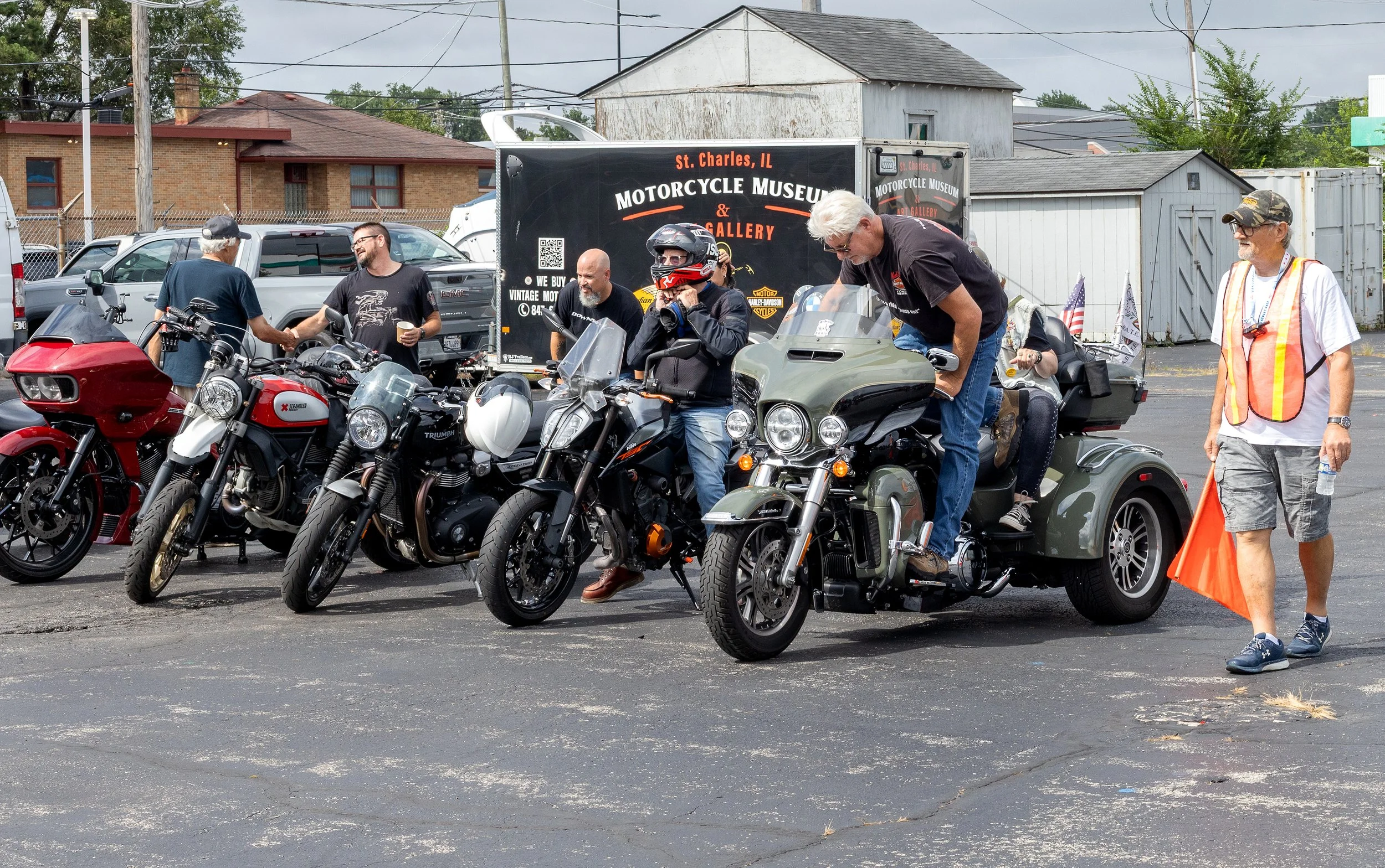 A group of people gathered around motorcycles at an outdoor event in a parking lot, with a backdrop of vehicles, a truck advertising a motorcycle museum, and small buildings. Some individuals are talking, one person is adjusting his motorcycle, and a