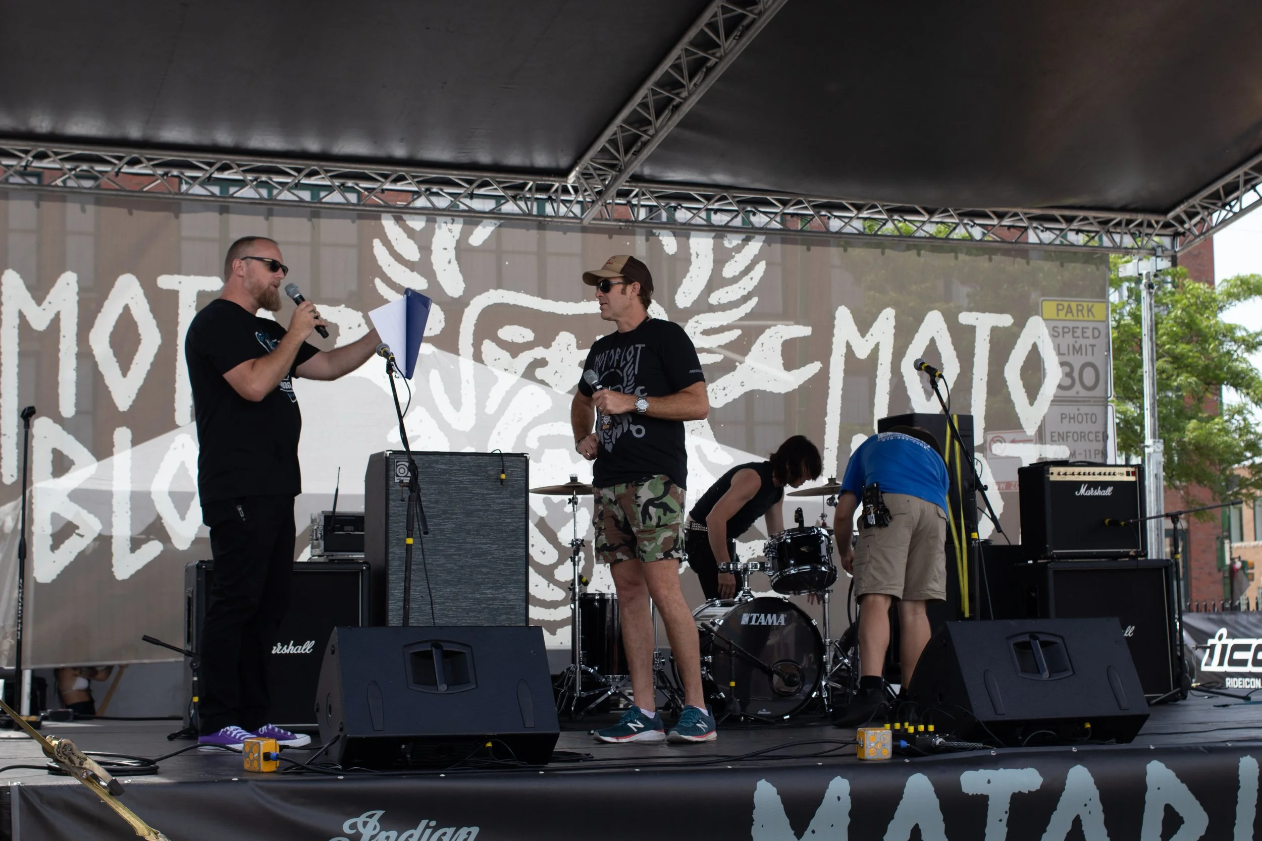 Two men are on an outdoor stage with musical equipment, one speaking into a microphone and the other listening. Several other people are preparing instruments, and a large banner with white graffiti-style art is in the background.