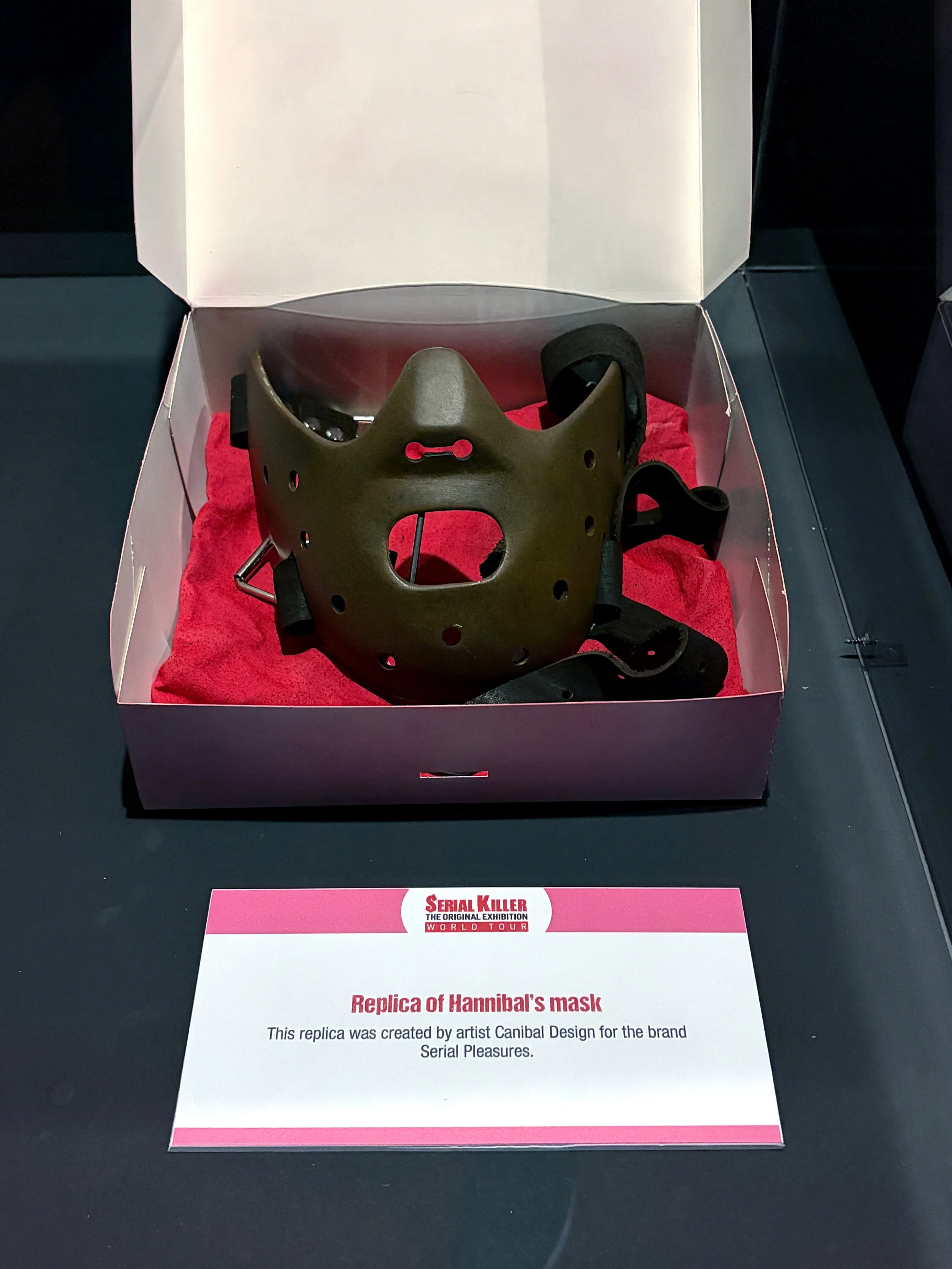 A replica of Hannibal's mask displayed in a box with a pink label that says 'Serial Killer The Original Exhibition World Tour' and an informational card describing the replica at the bottom.