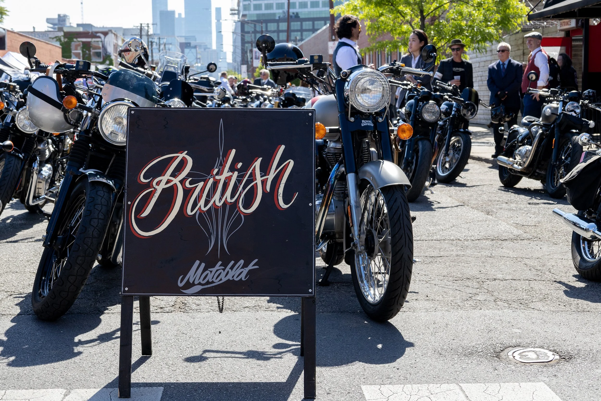 Row of motorcycles parked outdoors with a sign in front that reads 'Buffle' and some people standing in the background, including a man in a suit and a woman in a white shirt with dark vest, in an urban setting with trees and buildings.