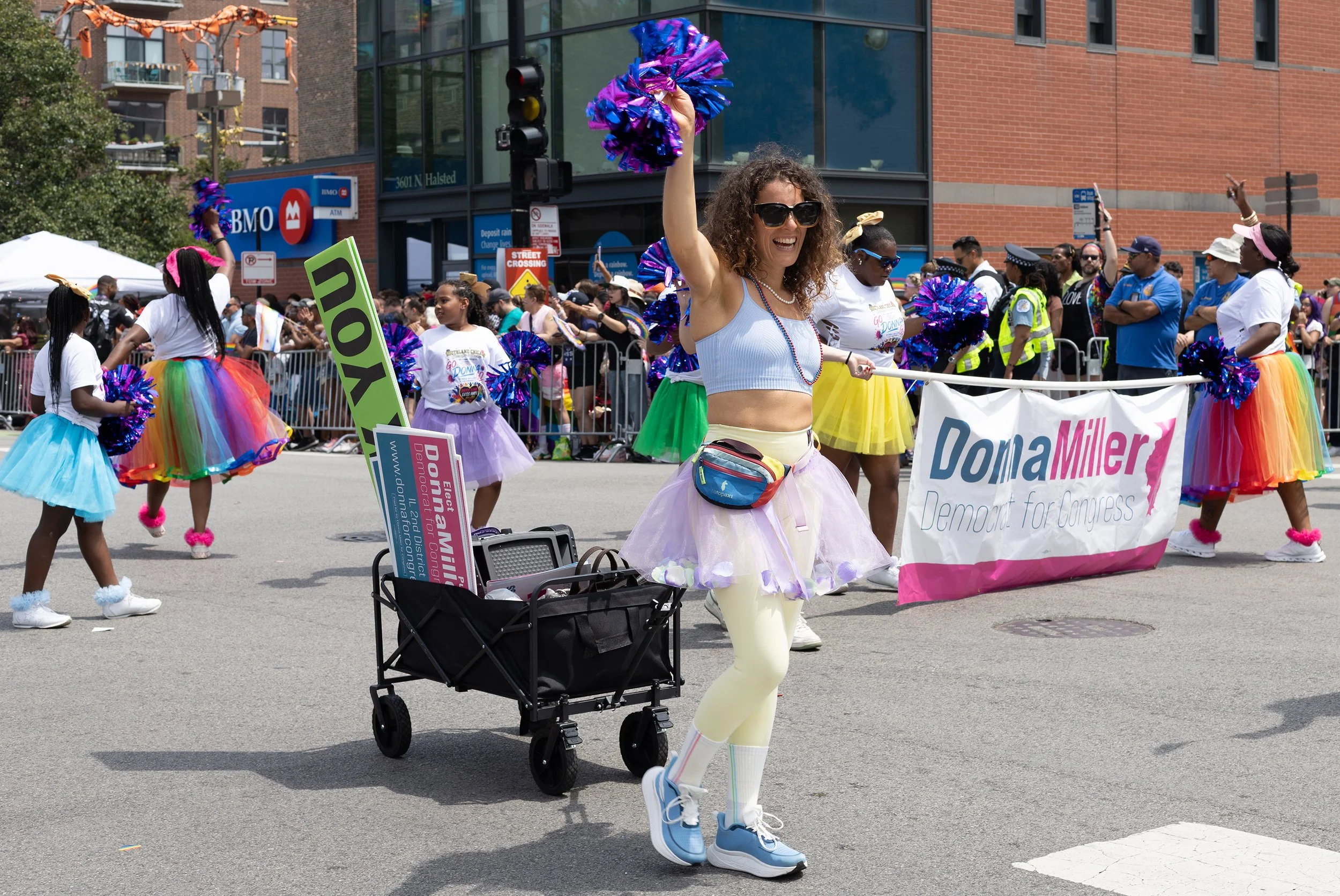 People participating in a parade, with women wearing colorful tutus and holding blue metallic pom-poms. A woman in the foreground is smiling, wearing sunglasses, a white crop top, yellow pants, and sneakers, with a stroller nearby. There are marchers