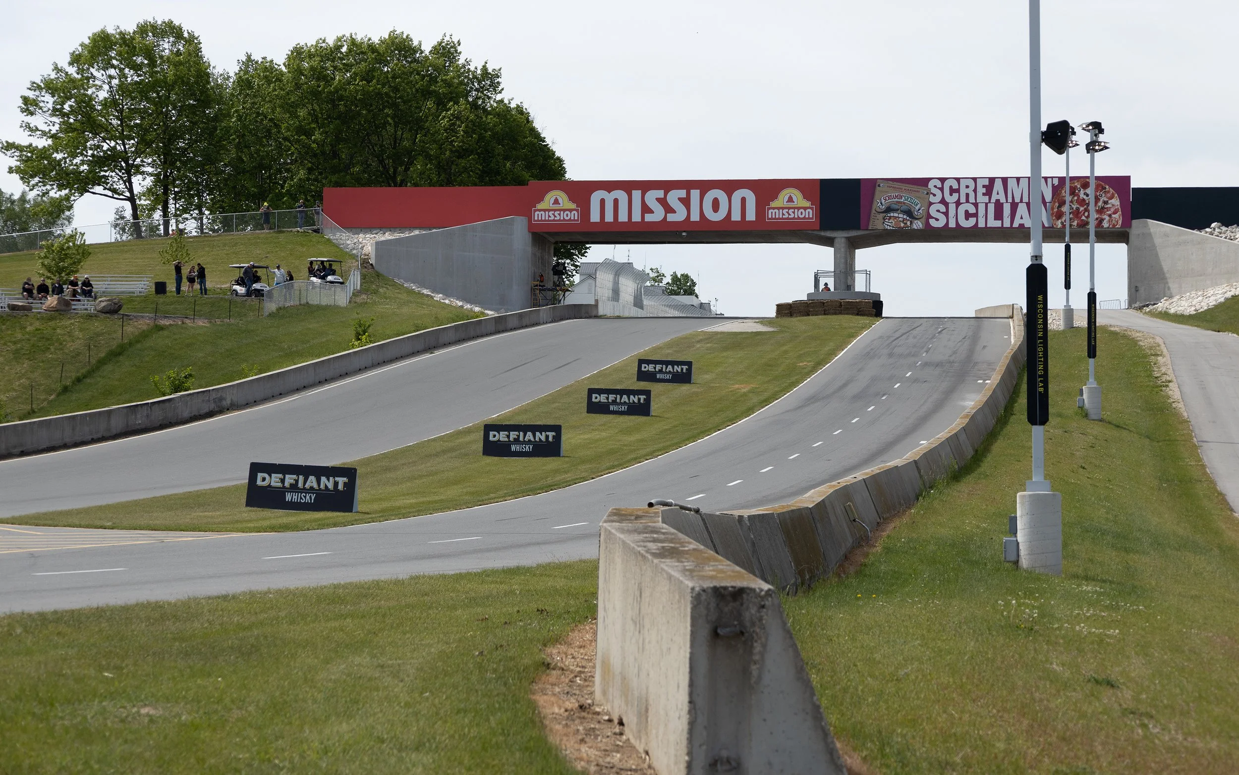 A race track with a bridge over it displaying advertisements and sponsors, with grassy hills, trees in the background, and light poles on the side.