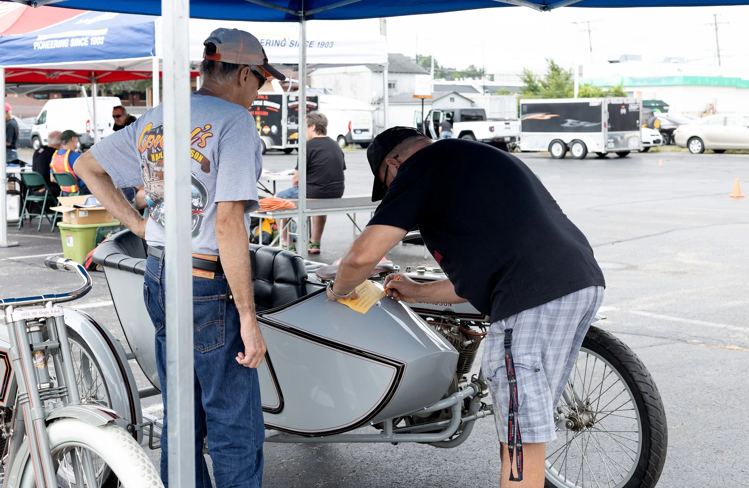Two men working on a drag racing motorcycle at an outdoor event under a blue canopy, with numerous trailers and cars parked in the background.