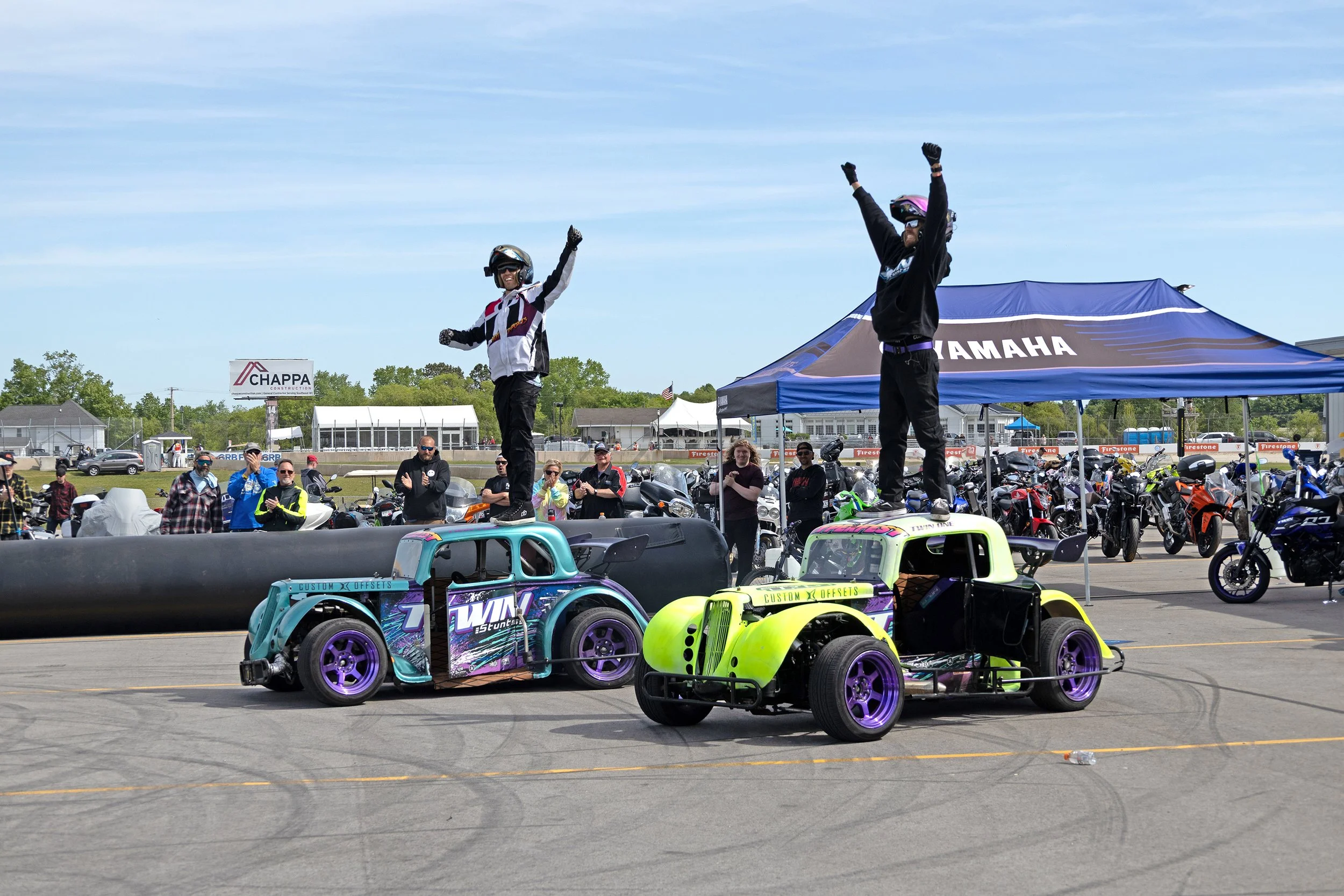 Two people standing on cars at a racing event, with one raising their hands in celebration. There are motorcycles and a crowd in the background under a blue canopy, with a clear sky overhead.