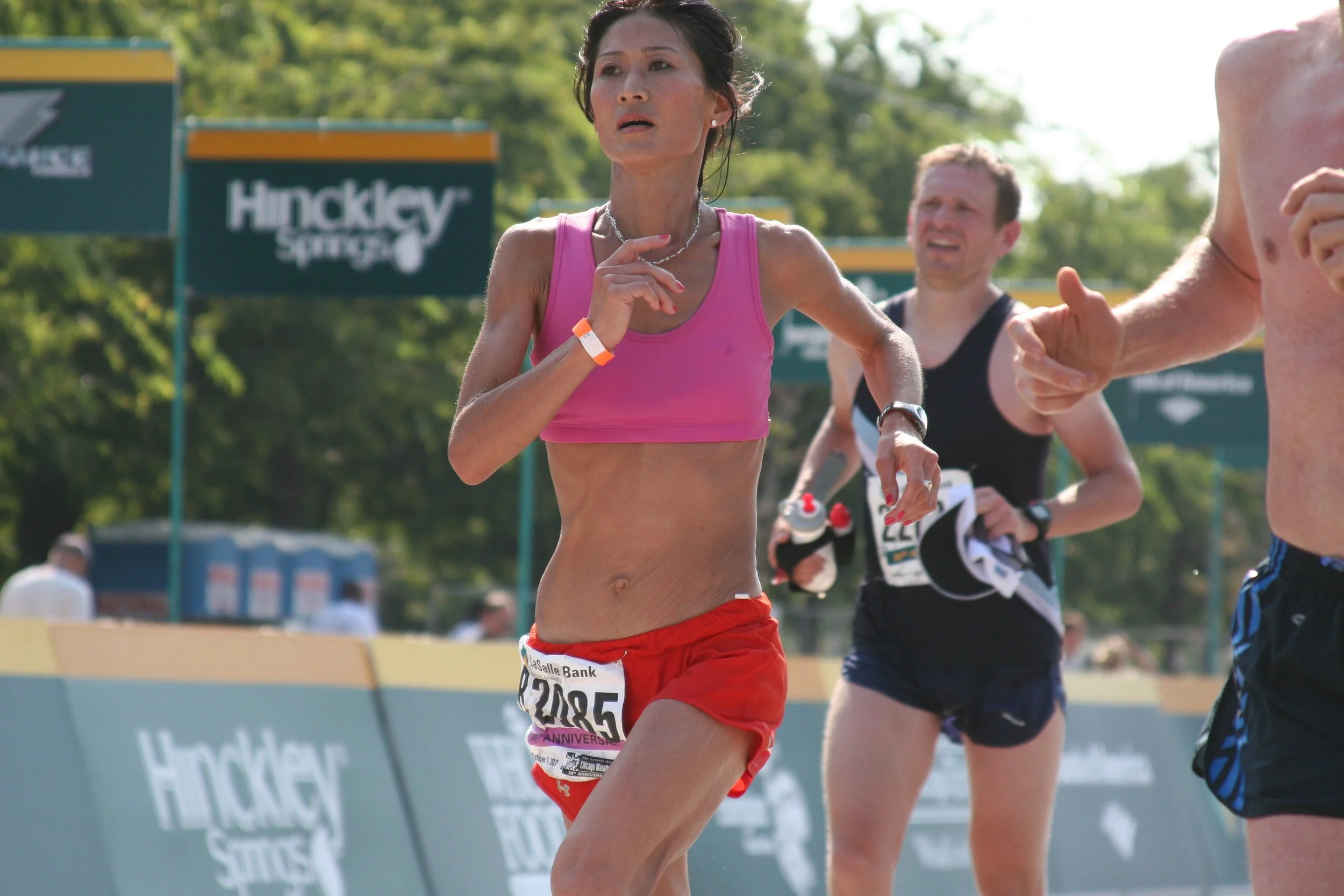A female runner in a pink sports bra and red shorts competing in a marathon, with other runners and a sign reading "Hockey" in the background.