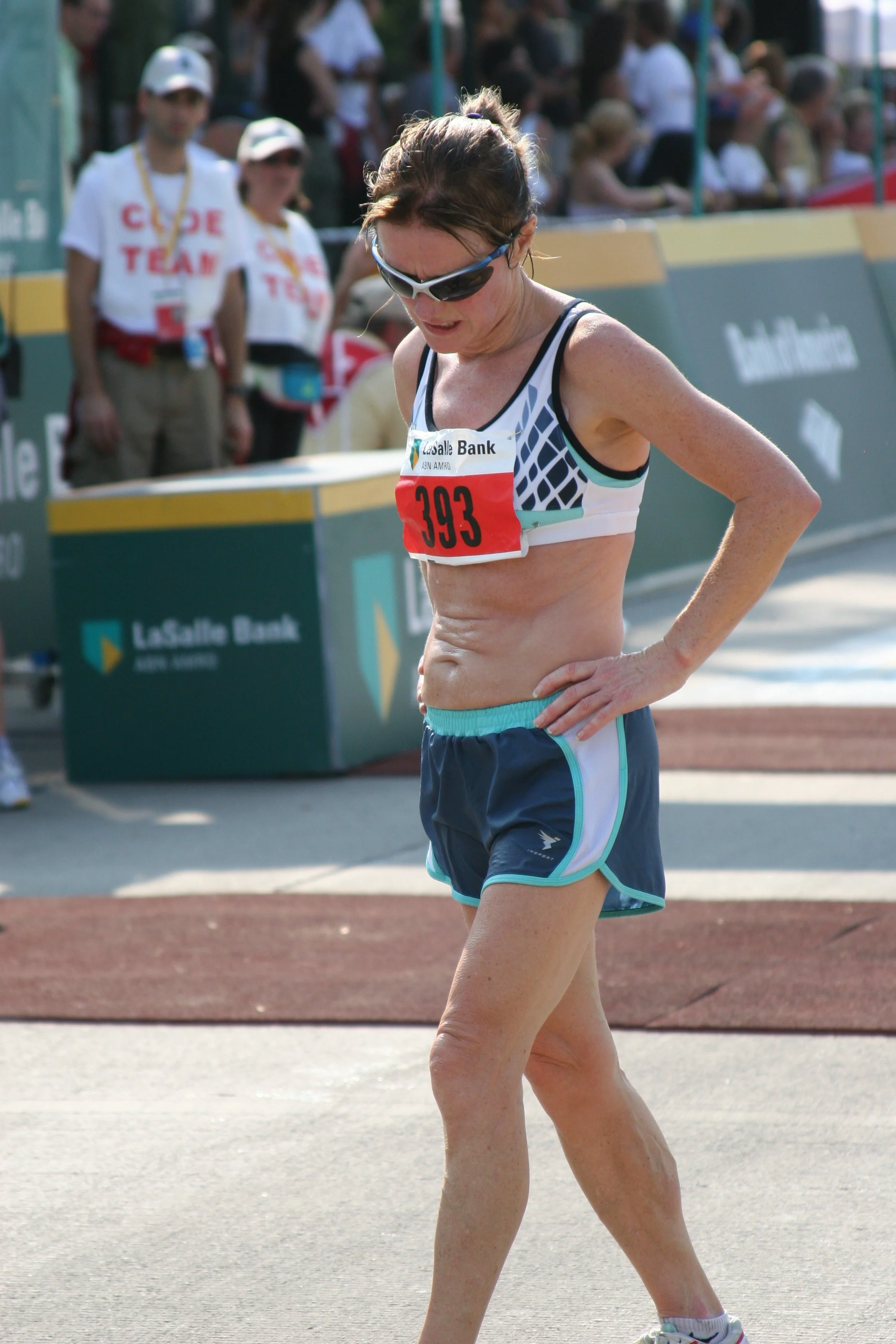 A female athlete in running gear, wearing sunglasses and a bib number 393, looks down during a race, with spectators and event staff in the background.