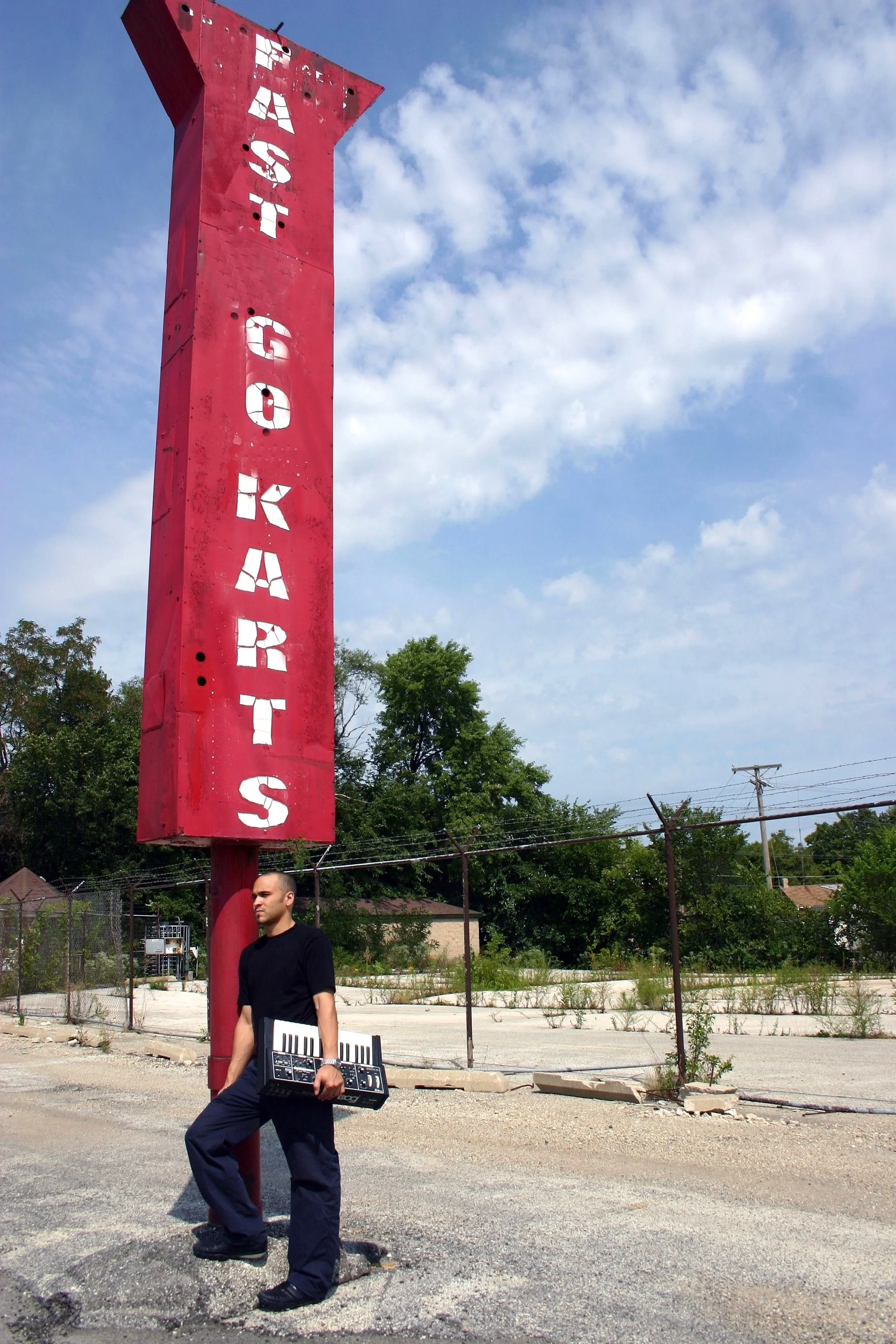 A man holding a keyboard and a synthesizer stands next to a tall red sign with white letters spelling out "GAS STATION" against a blue sky with white clouds. There are trees, a fence, and a power line in the background.
