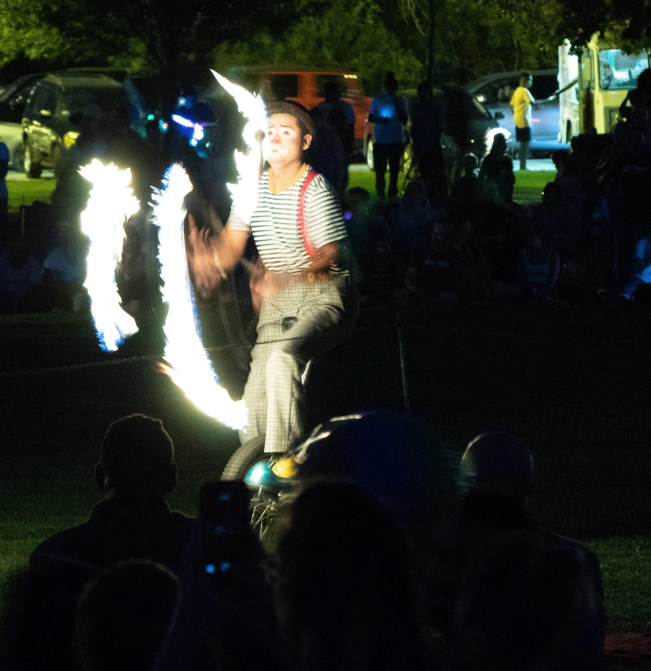 Nighttime scene with a performer in a fire show, holding fire props or eating fire, with an audience in the dark. Background features trees, cars, and a brightly lit food truck or street vendor.