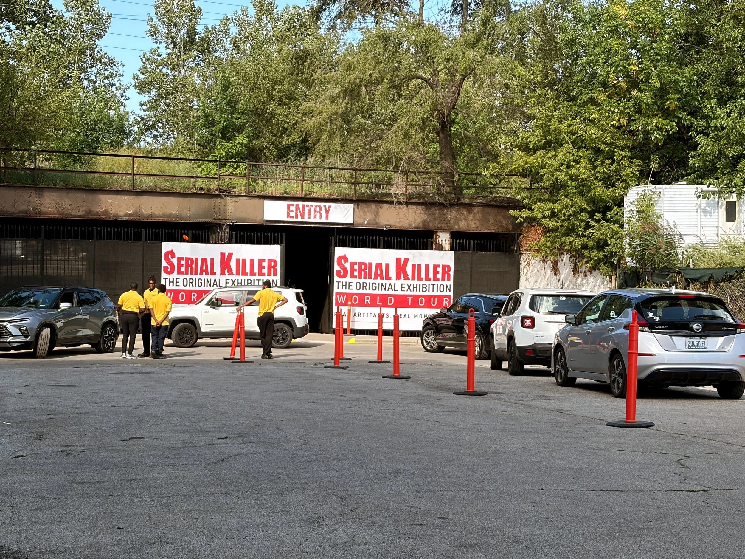 Parking lot with several cars and orange traffic cones in front. Large banners advertise the 'Serial Killer' exhibition on artifacts and real murder cases, with a white and red color scheme. In the background, trees and a building with an 'ENTRY' sig
