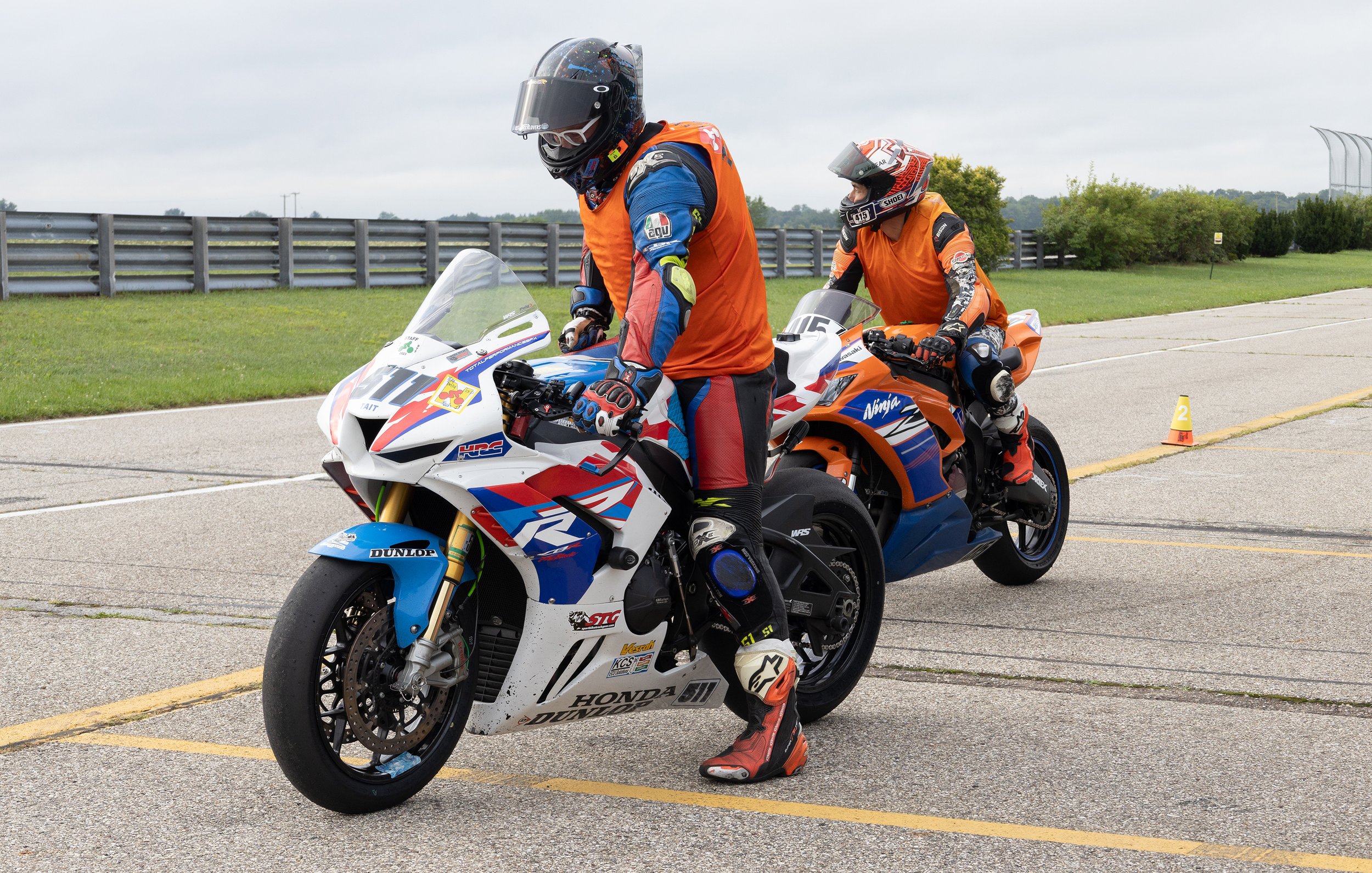 Two motorcycle racers in racing gear and helmets sitting on their racing bikes on a track, preparing for a race.