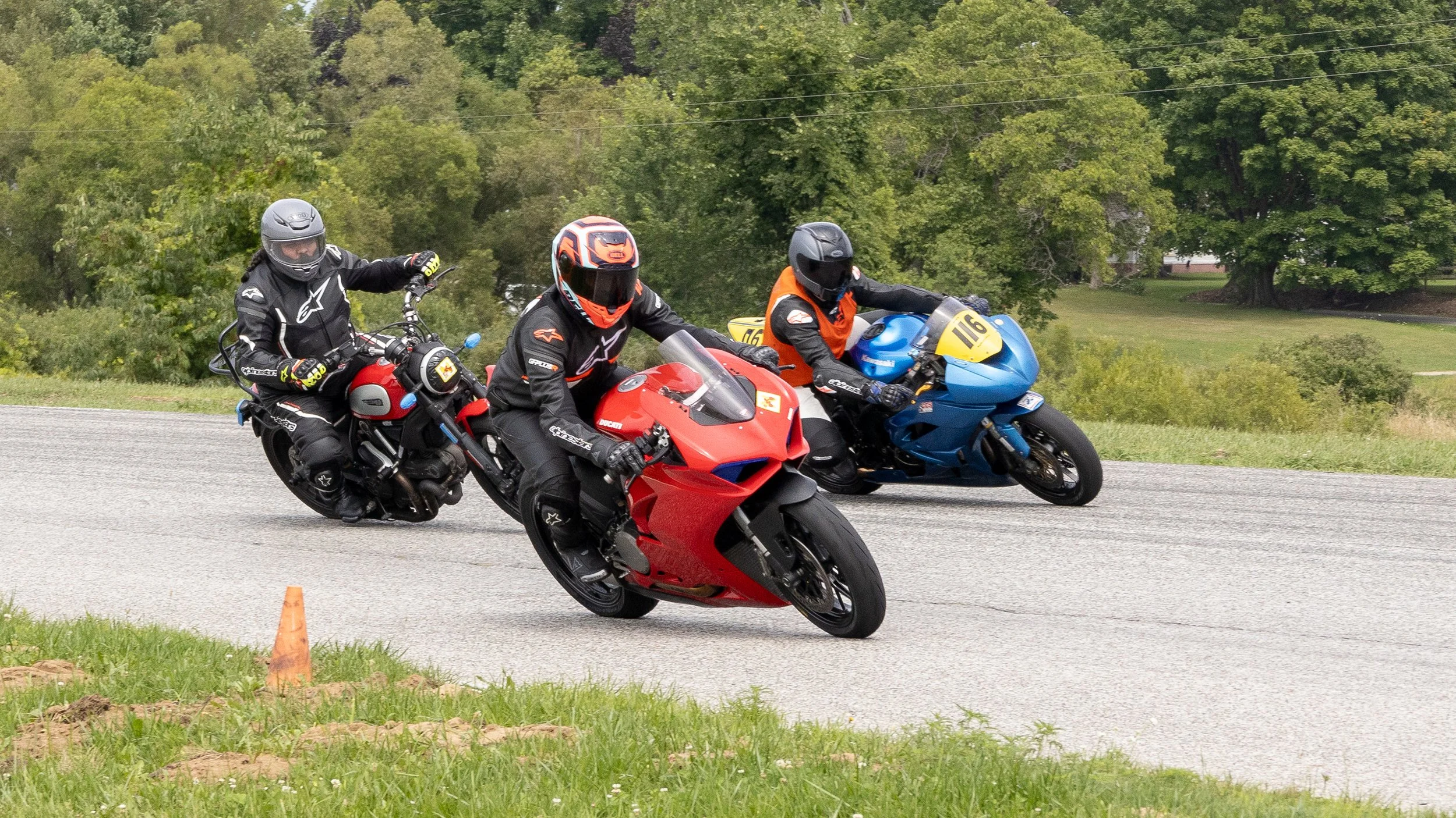 Three motorcycle racers navigating a turn on a race track, wearing full protective gear and helmets, with greenery and trees in the background.