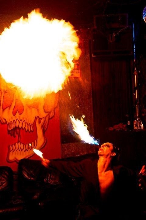 A fire performer is breathing fire in a dimly lit room with a mural of a skull with hearts on the wall behind.