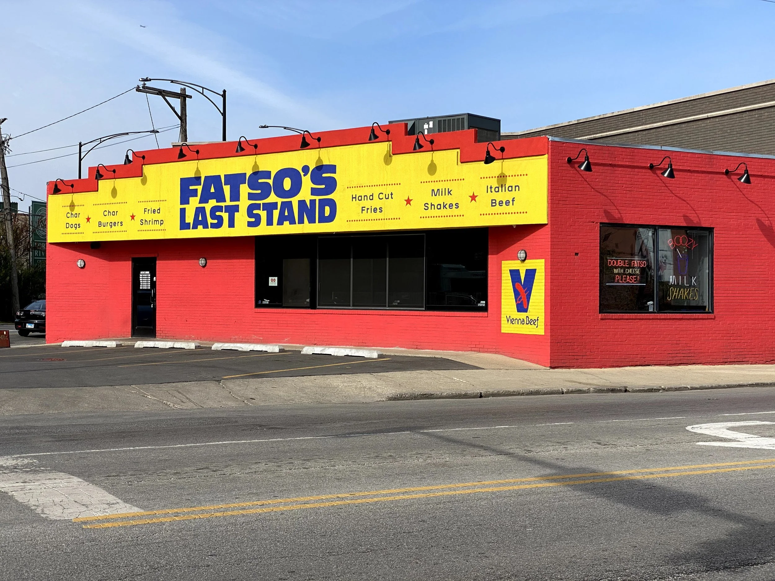 A red brick building with a large yellow sign that reads "Fatso's Last Stand" in blue letters, advertising char dogs, char burgers, fried shrimp, hand-cut fries, milkshakes, Italian beef, and Vienna beef. The building has a window with neon signs.