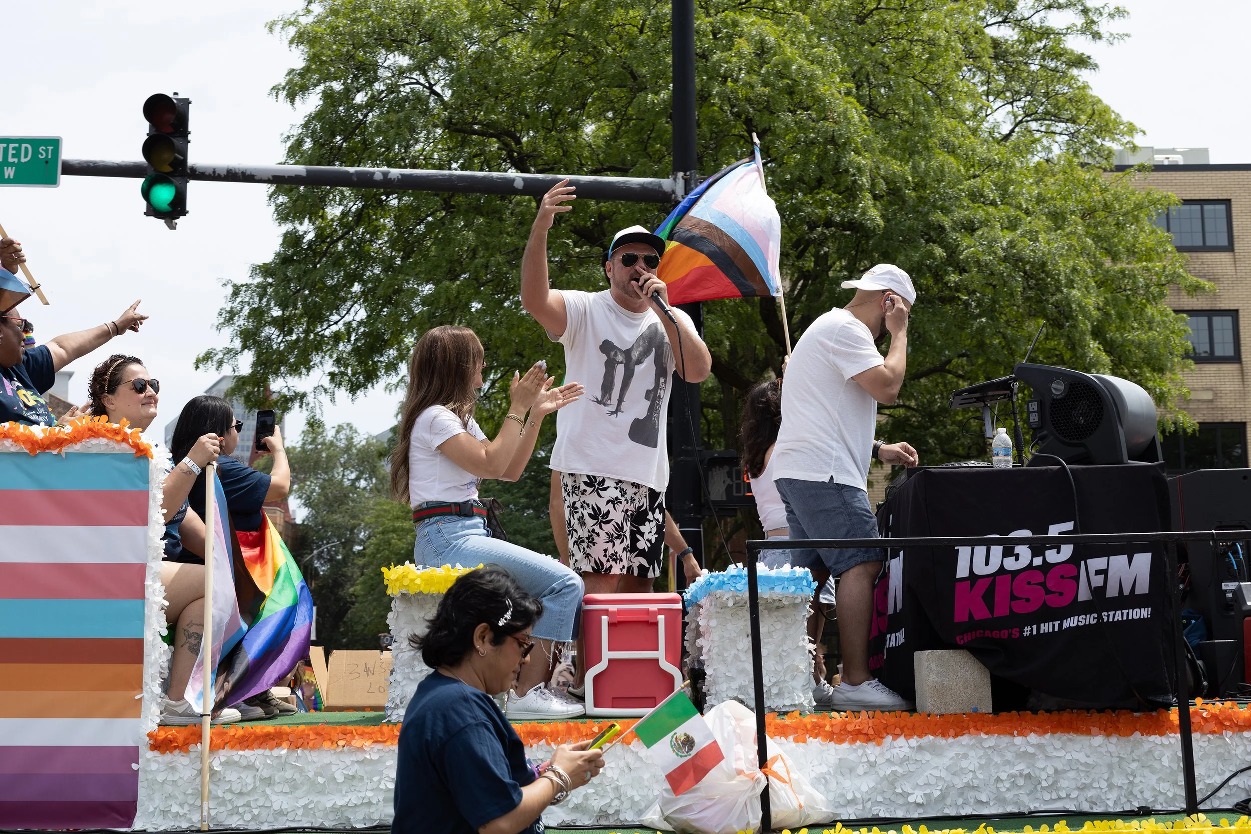A group of people celebrating at a pride parade, with a man speaking into a microphone on a float decorated with rainbow flags and floral arrangements, people waving flags, some wearing white hats and sunglasses, and a large rainbow LGBTQ+ flag in th