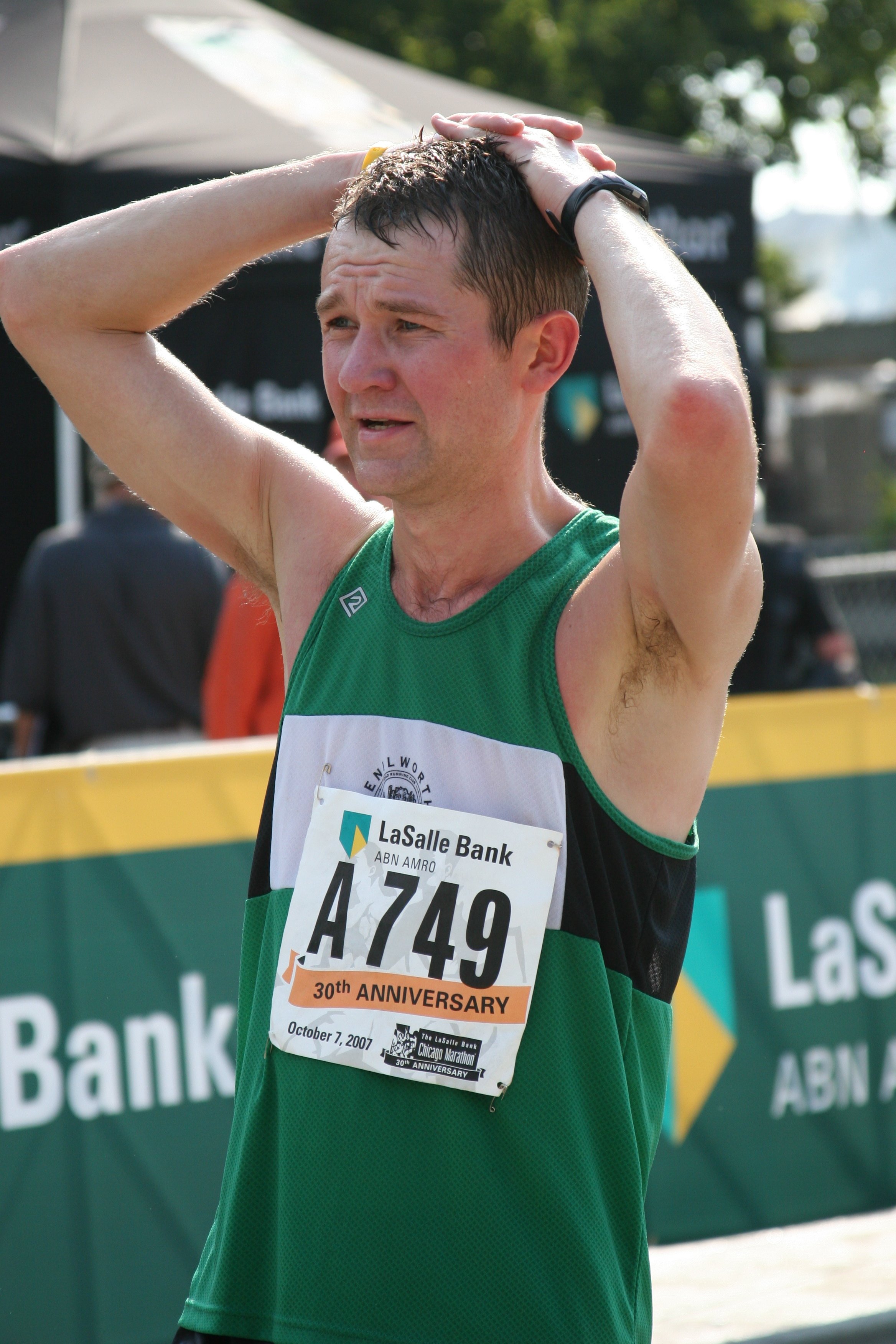 A male marathon runner in a green and black shirt with a race bib and a stopwatch on his wrist, wiping his face after finishing a race outdoors.
