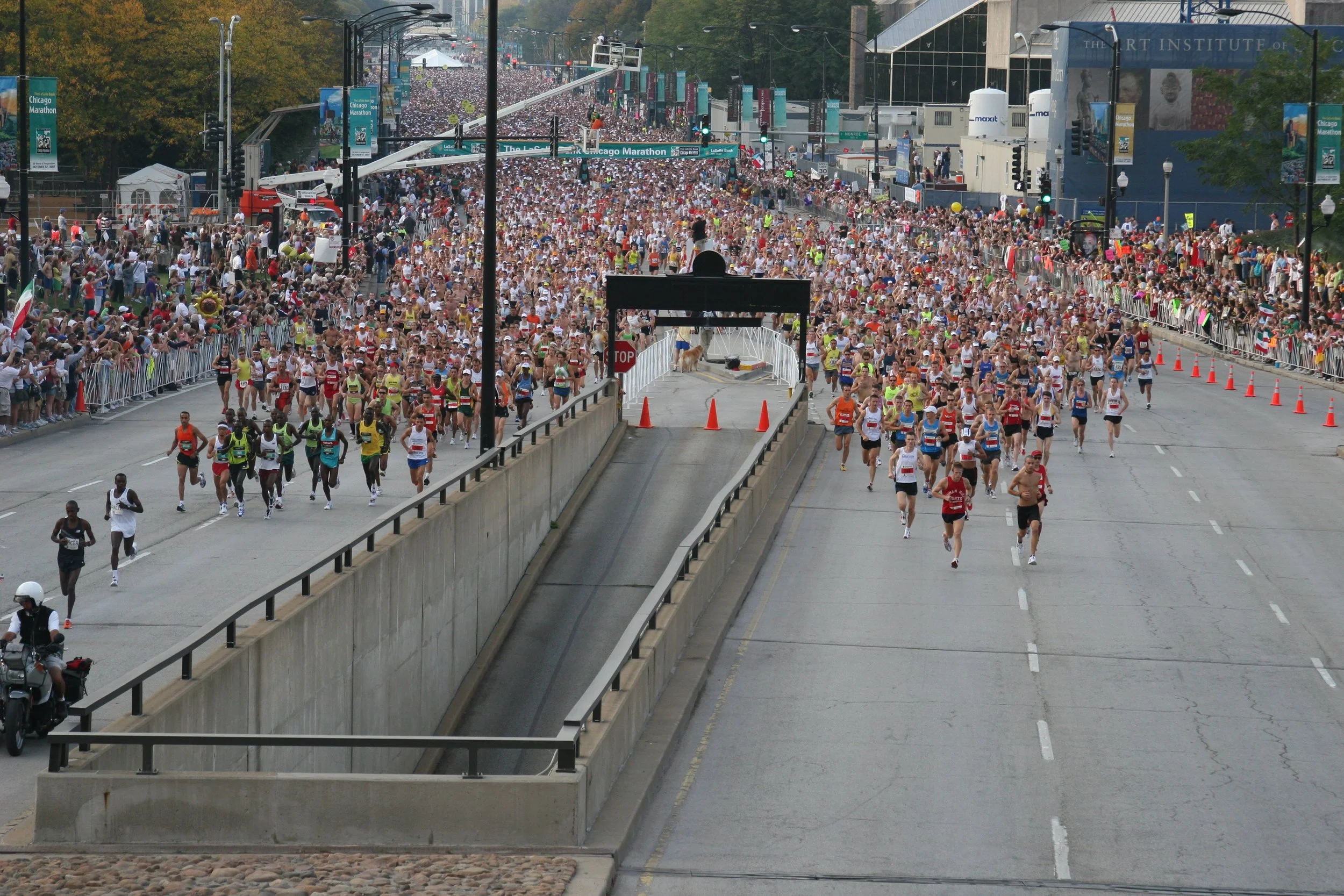 Crowd of marathon runners and spectators at the Chicago Marathon, with banners and a large crowd in the background, as seen from a street viewpoint.