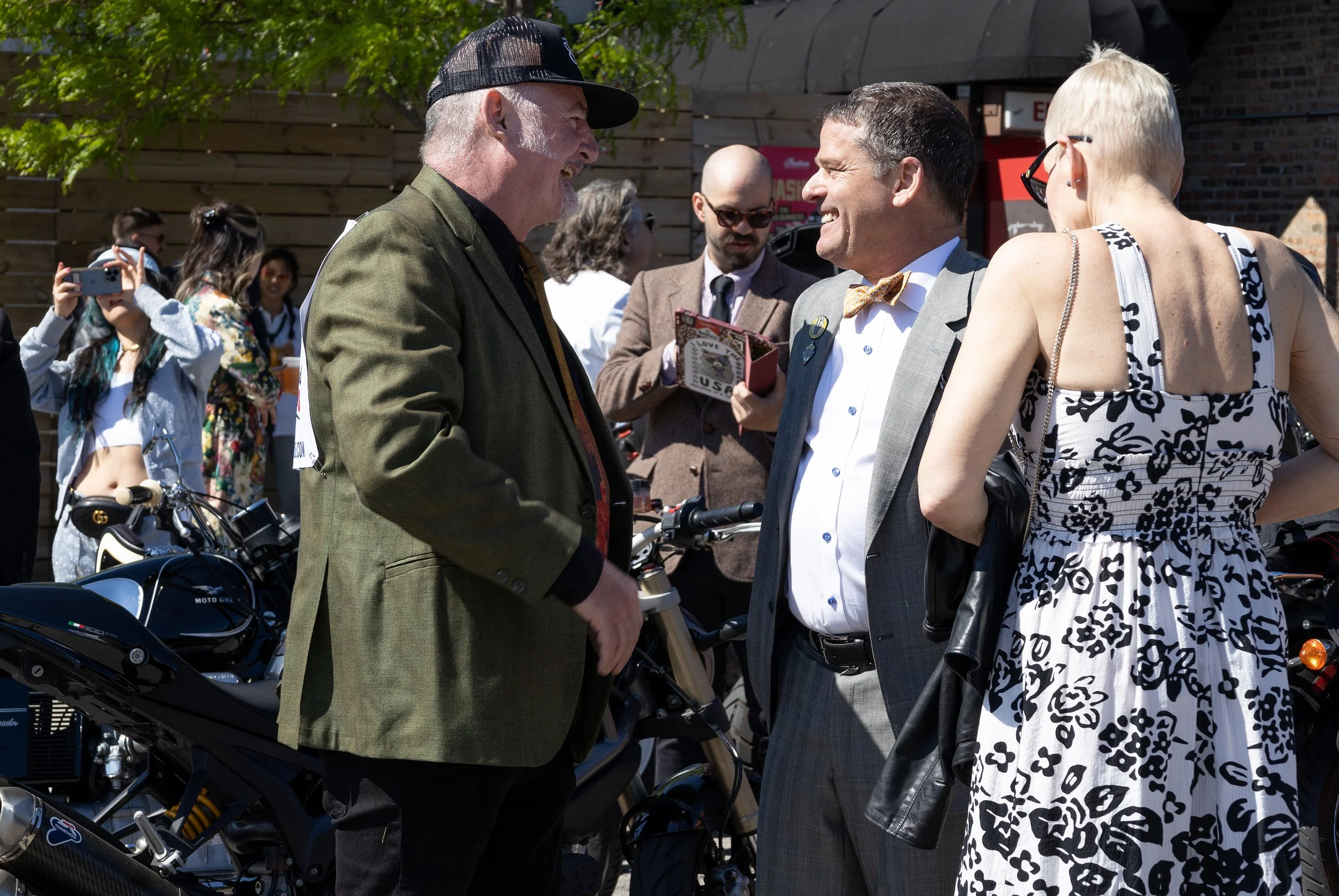 Three people are engaged in a conversation. The man on the left has a gray beard, is wearing a dark cap, white shirt, and a green blazer. The man in the middle has dark hair, is smiling, dressed in a light gray suit with a bow tie. The woman on the r