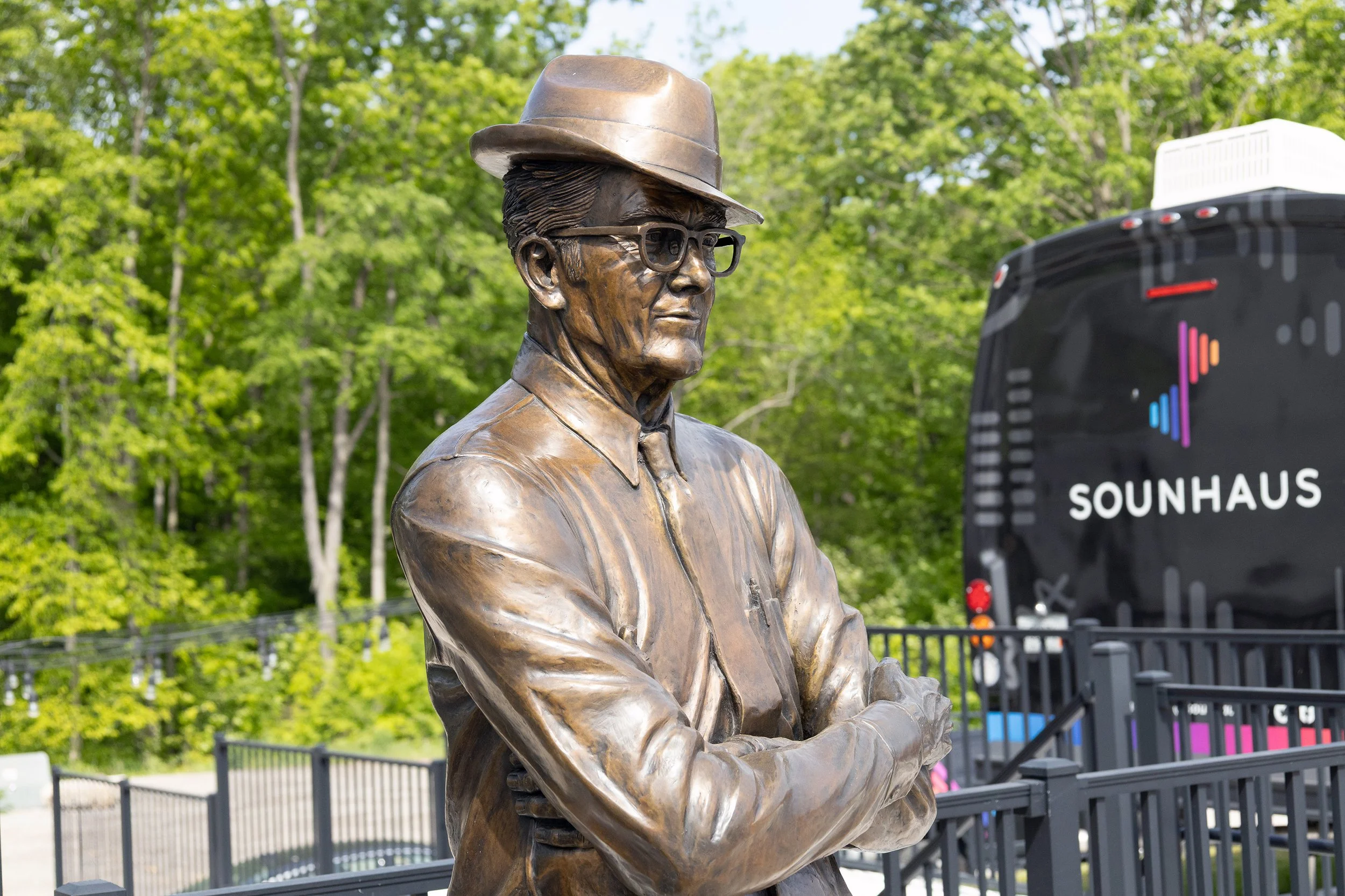 Bronze statue of a man wearing glasses, a suit, and a fedora hat, with a backdrop of green trees and a sound equipment truck.