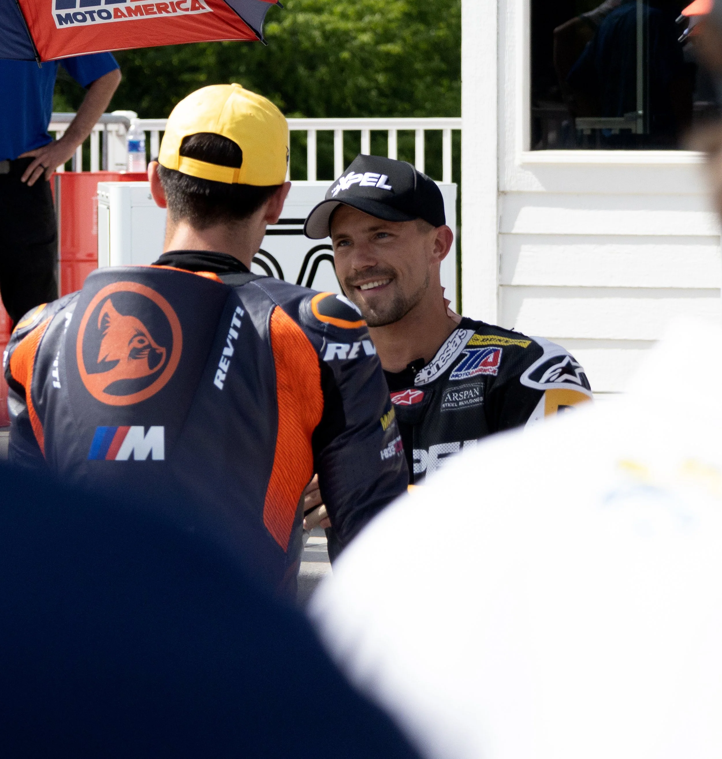Two race car drivers in racing suits and caps, engaged in conversation, with a third person holding an umbrella with the "MotoAmerica" logo, in an outdoor setting during daytime.