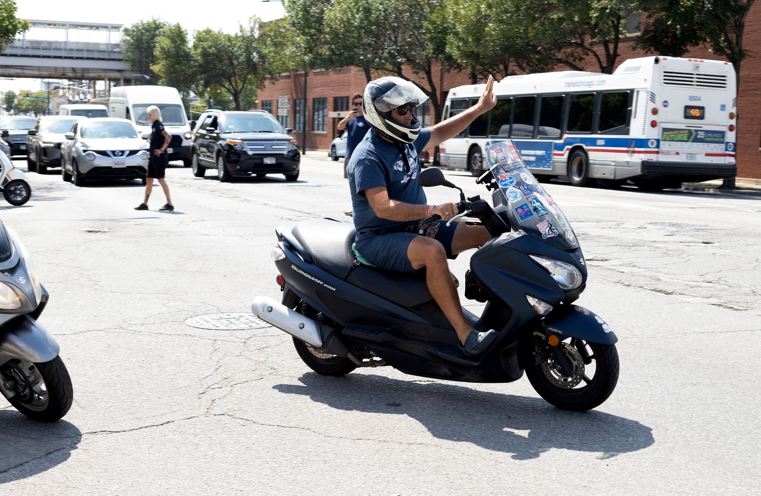 A man wearing a helmet rides a black scooter across a busy city street. There are cars and a bus in the background, as well as trees and brick buildings lining the street.