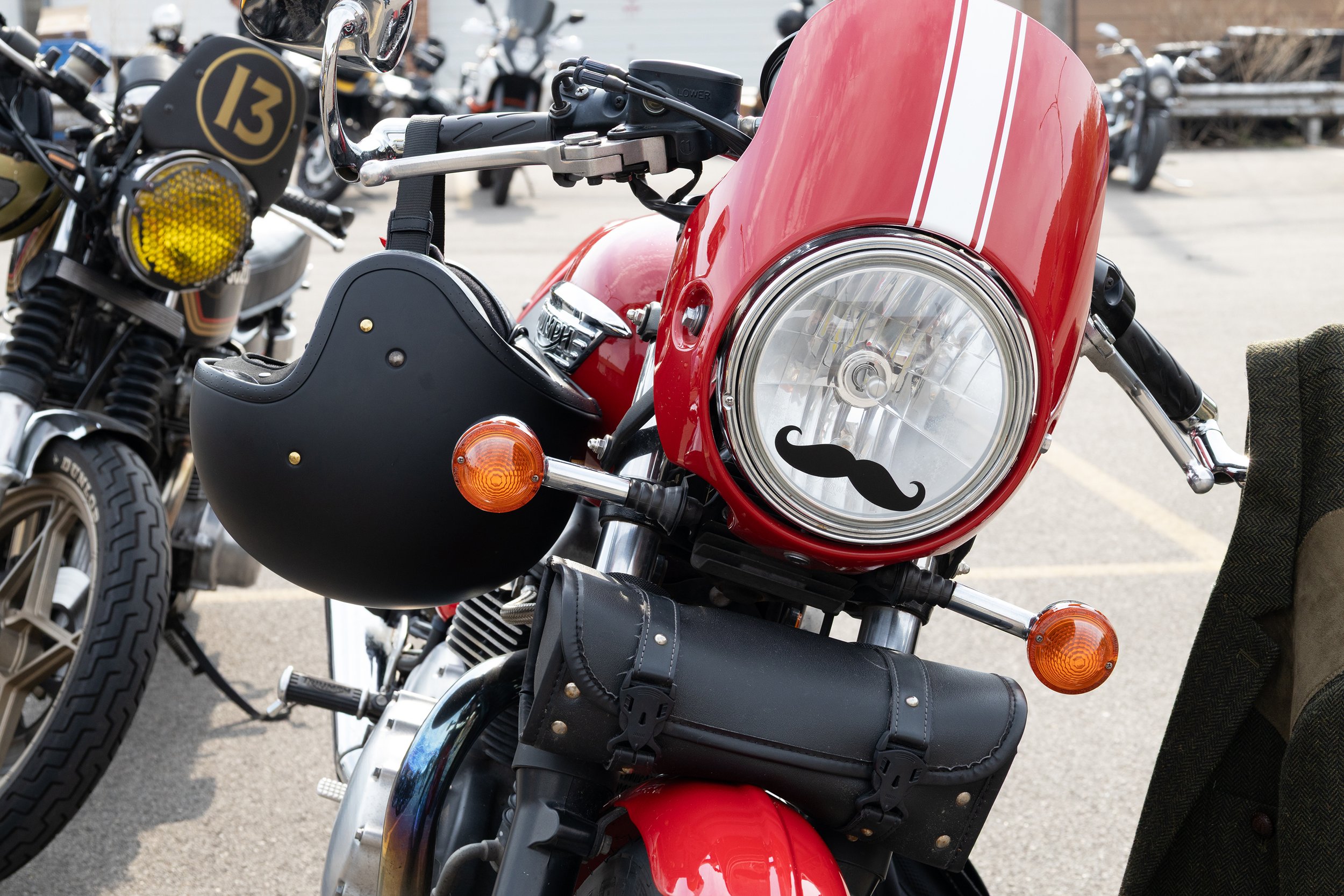 Close-up of a red motorcycle with a large round headlight and a license plate with a mustache graphic, parked among other motorcycles.