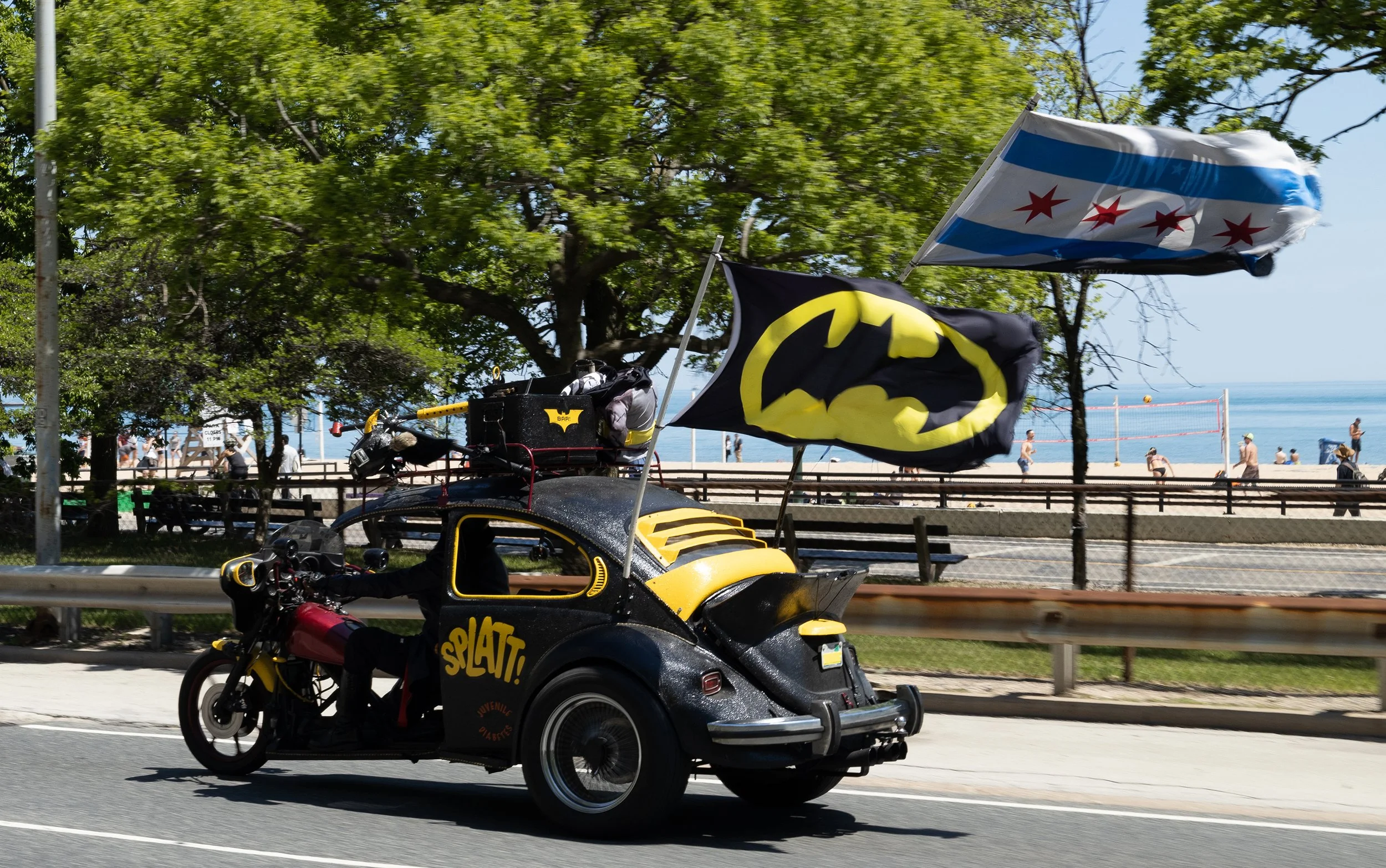 A yellow and black pedal-powered vehicle called a 'splat' with flags of Chicago and Batman logos, driving on a street near a beach, with trees, people, and volleyball courts in the background.