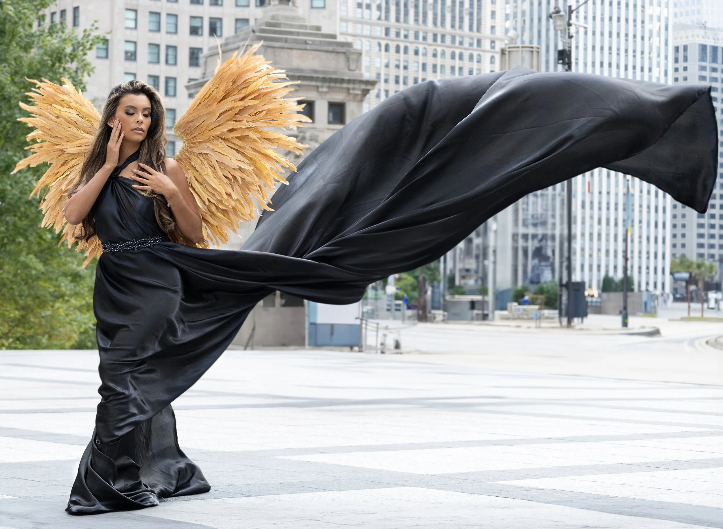 A woman with angel wings and long brown hair performing a dance move in an urban park, wearing a black dress and black shoes, with city buildings in the background.