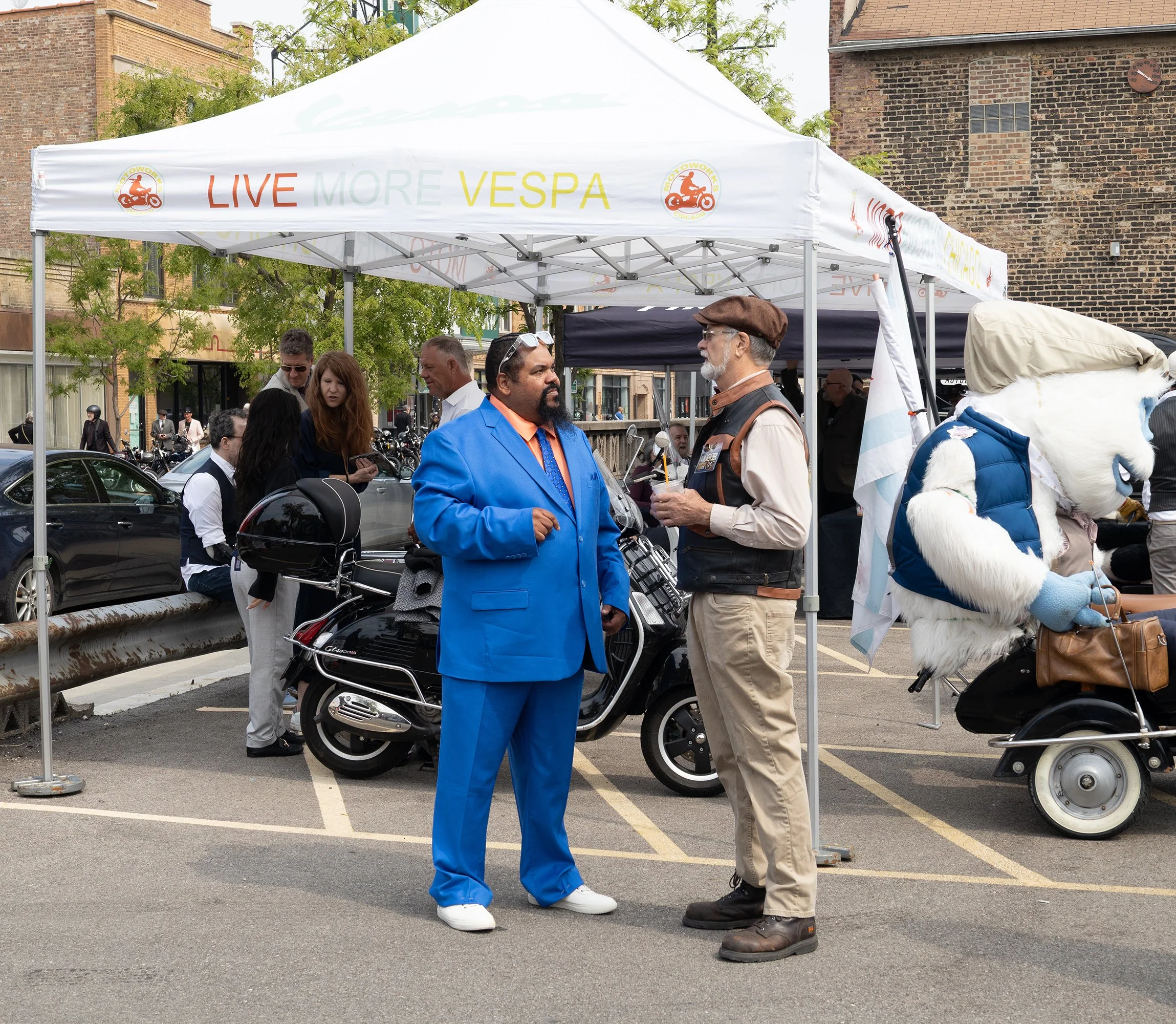 People gathered under a white tent that has a sign reading 'Live More Vespa' with a small motorcycle logo, in an outdoor urban setting with brick buildings and trees, some individuals talking, a person with a blue suit, and a person in a vest and cap