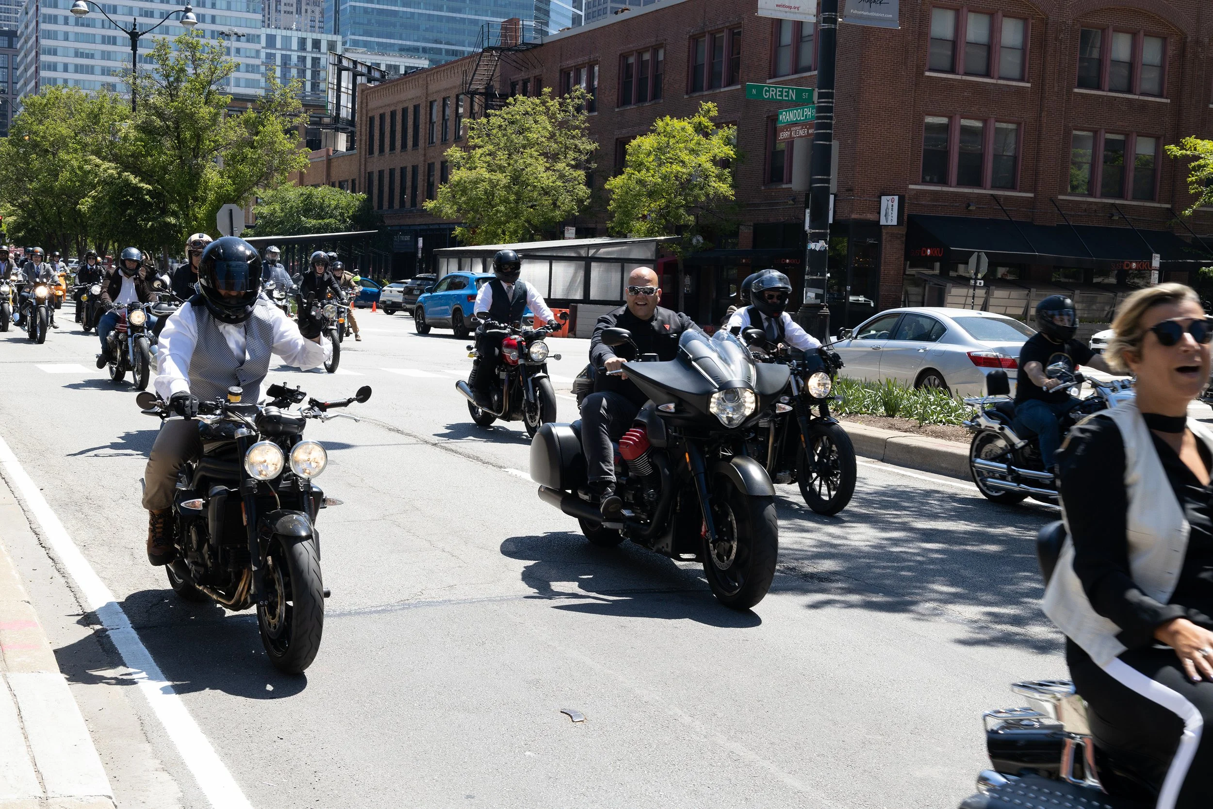 A group of motorcyclists riding on an urban street intersection in daylight, with trees, cars, brick buildings, and modern high-rise buildings in the background.
