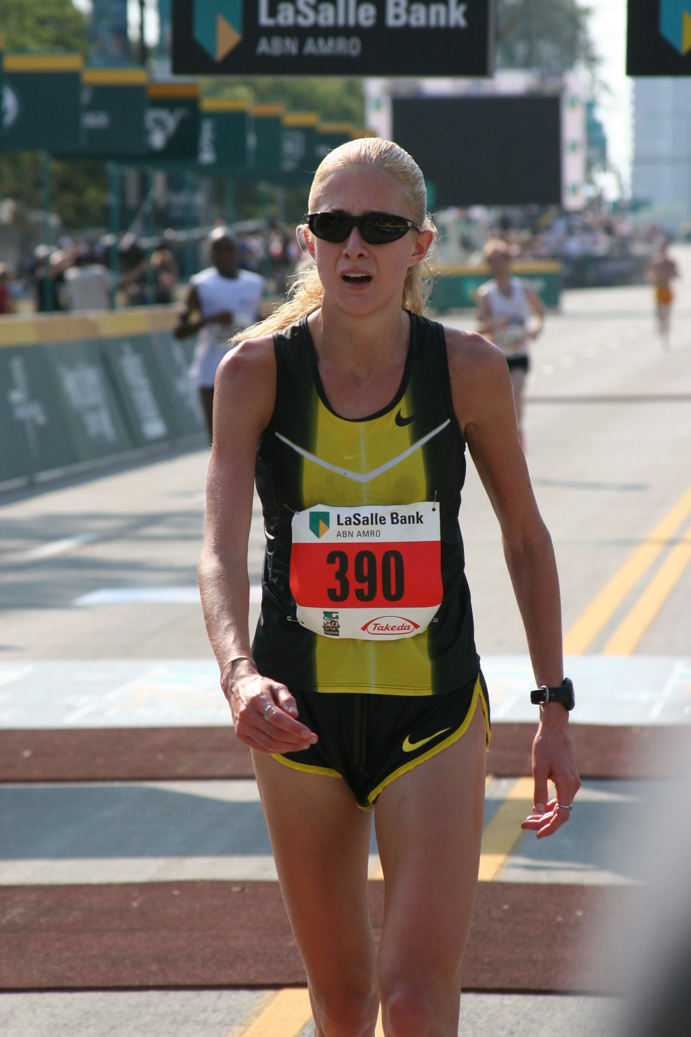 A female marathon runner wearing sunglasses and a yellow and black Nike race outfit, crossing the finish line with a focused expression. There are other runners and a banner in the background that says 'LaSalle Bank'.
