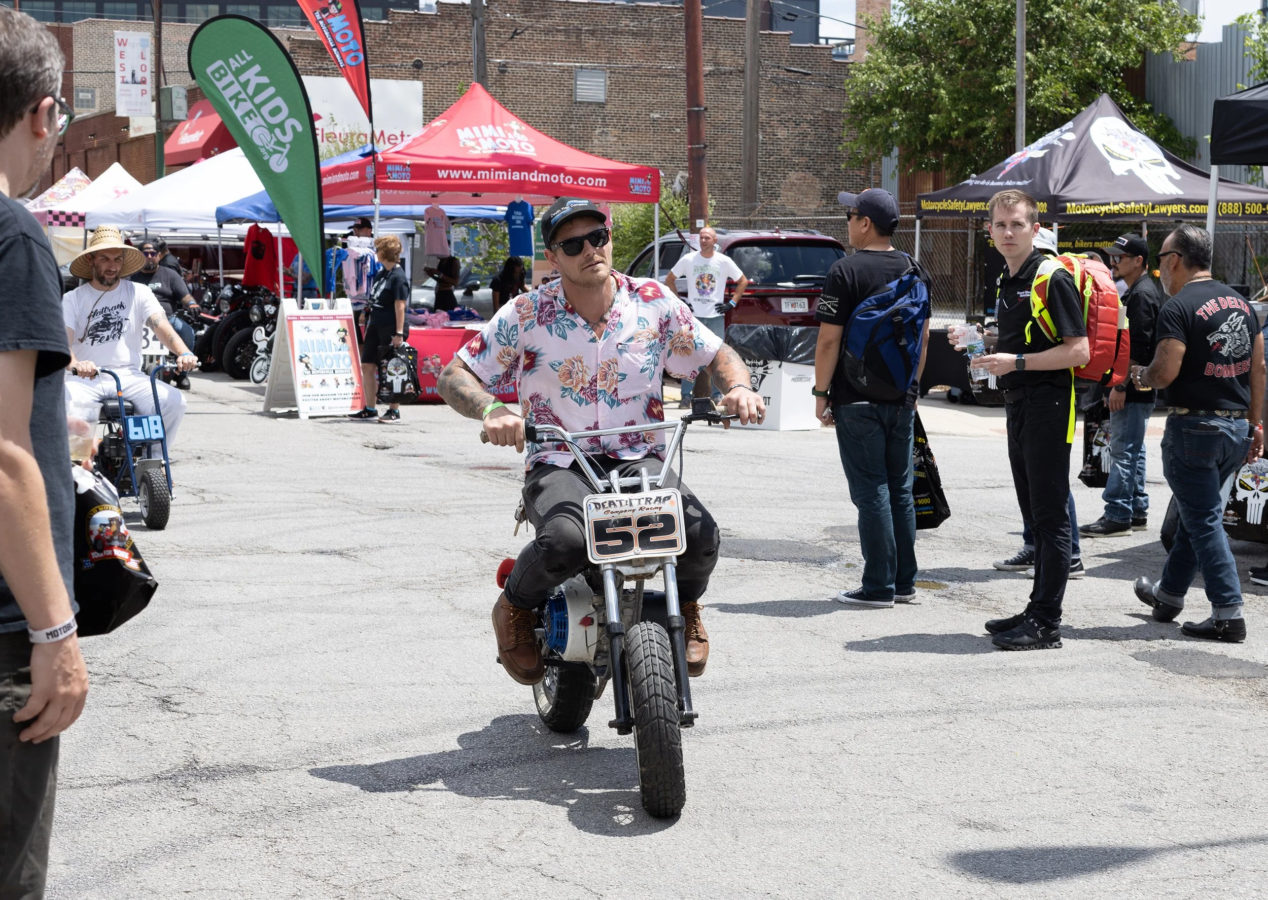 A man in a floral shirt and sunglasses riding a motorcycle with a race number 52 in an outdoor event, surrounded by people, tents, and banners.